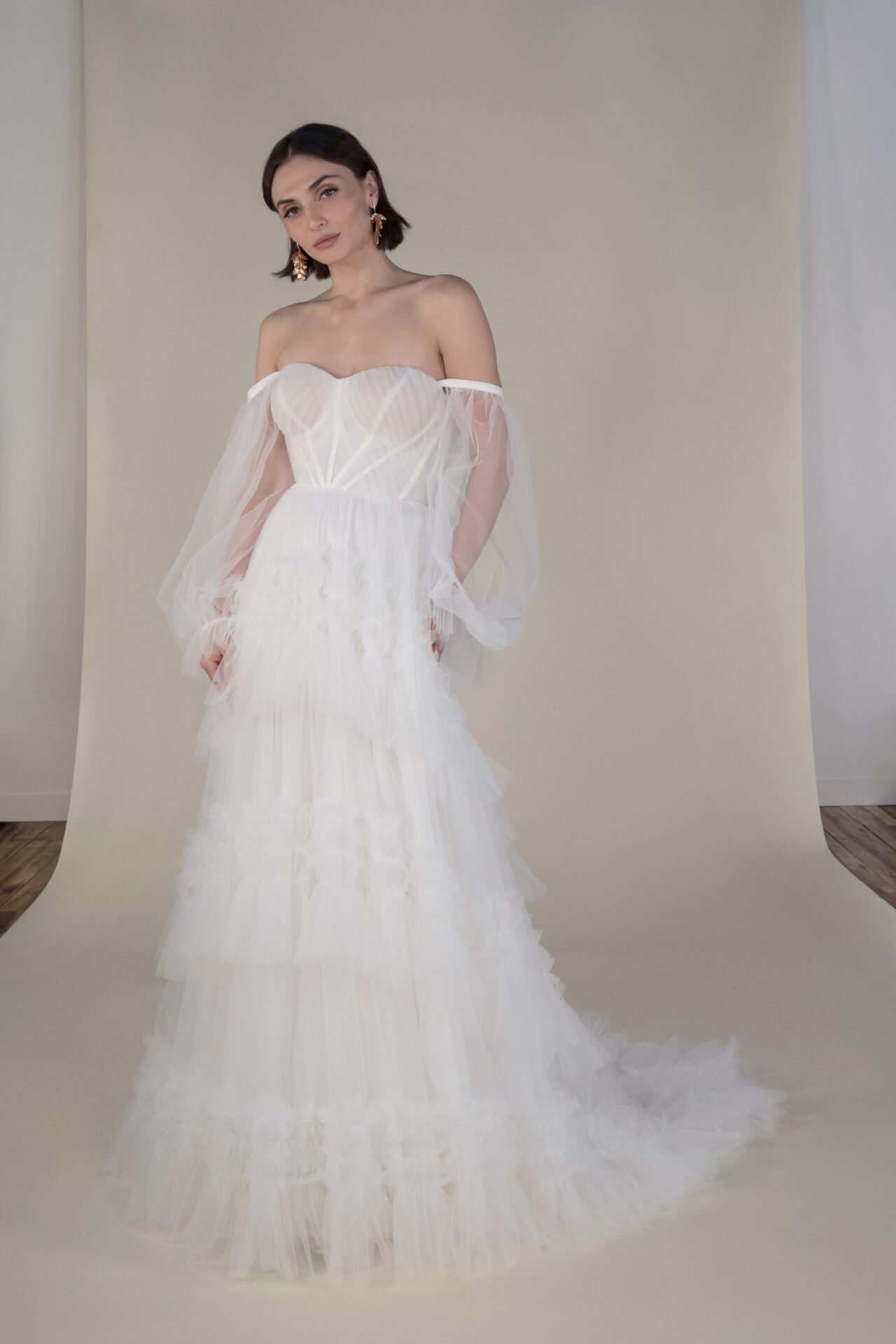 Woman in a white off-shoulder wedding dress with sheer puffed sleeves, standing on a light-colored backdrop in a room with wooden flooring.