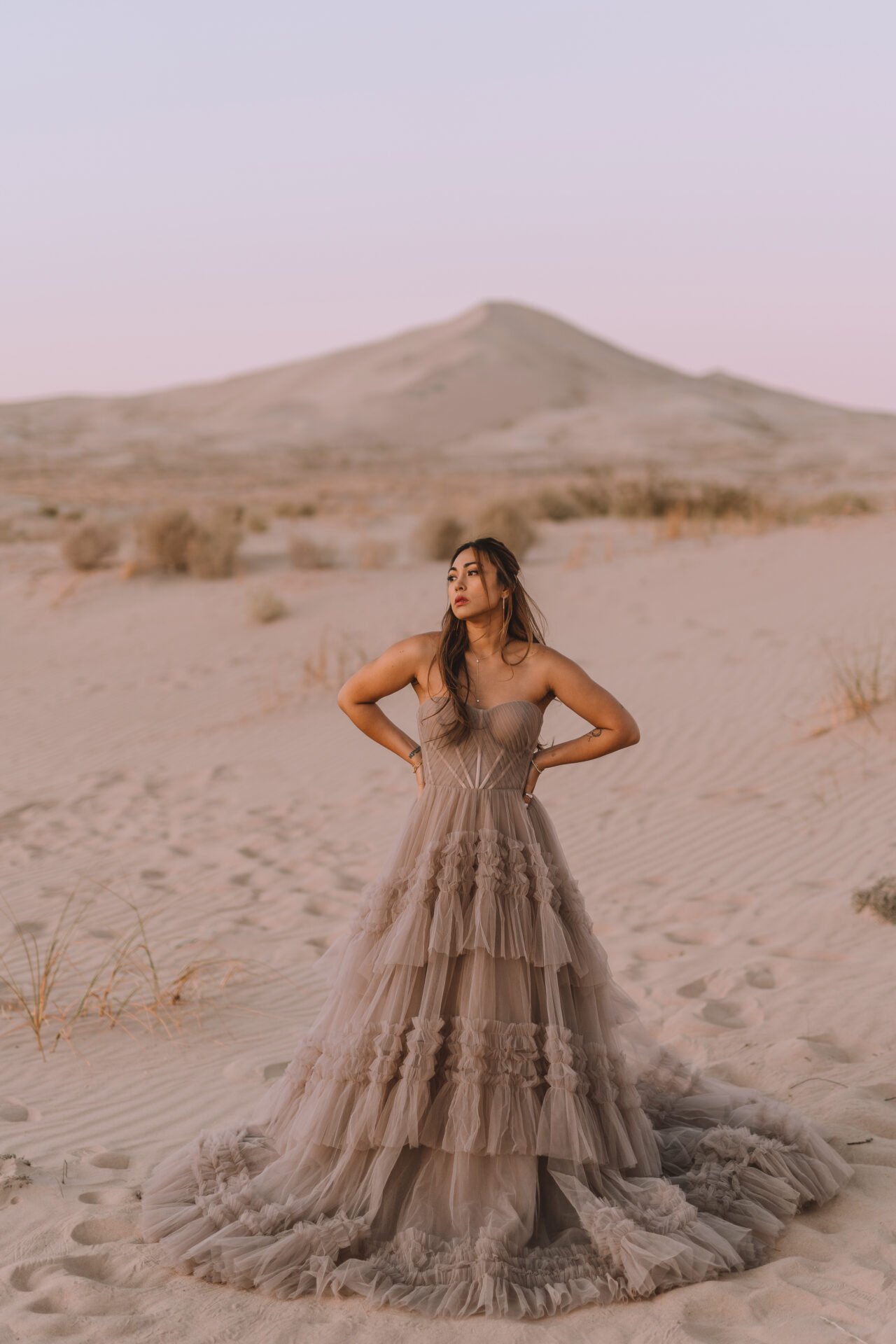 A woman wearing a strapless beige tulle gown with ruffles stands in a sandy desert landscape during sunset or sunrise.