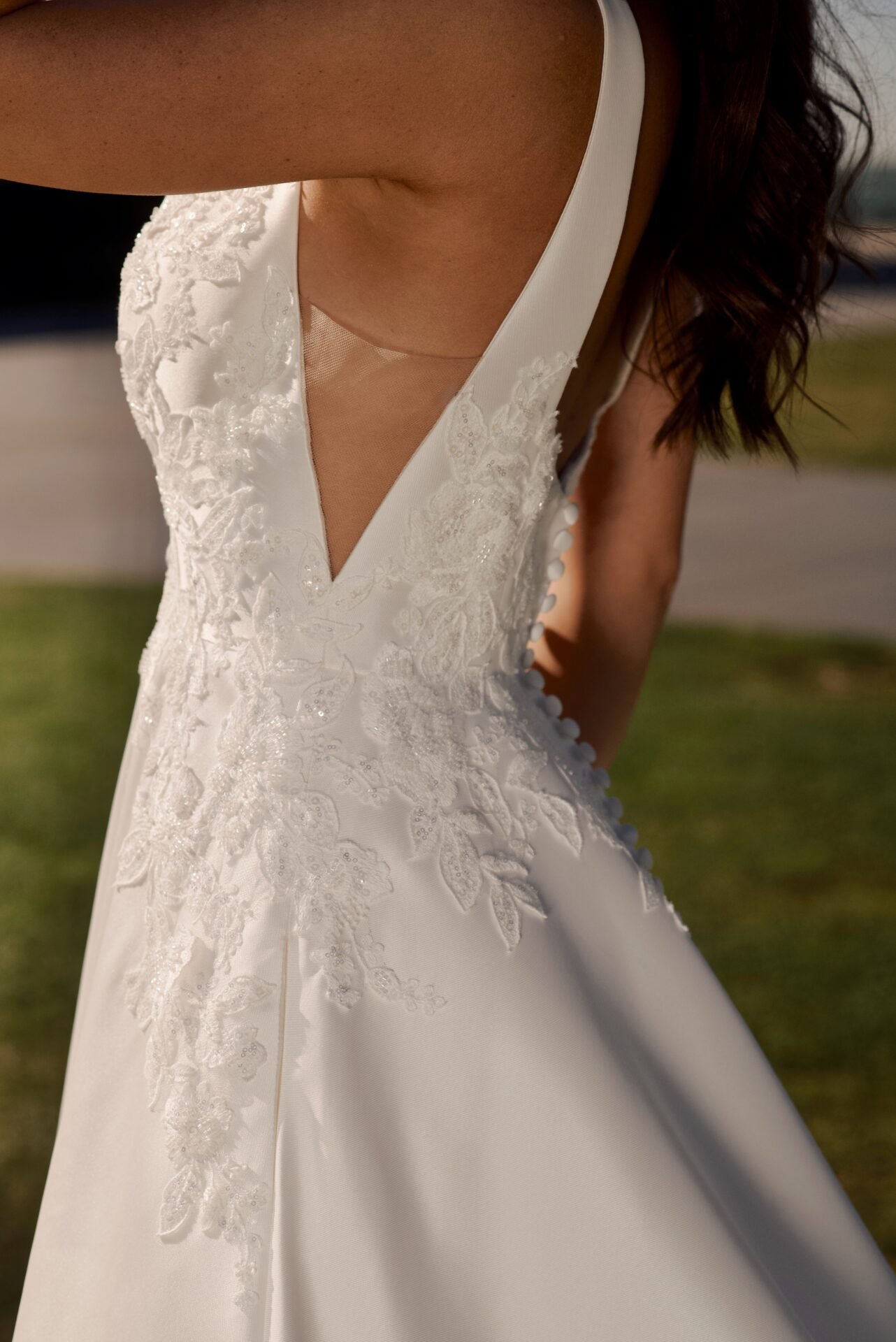Close-up of a bride's wedding dress with lace appliqué, featuring a deep V neckline and buttons on the side, outdoors on a sunny day.