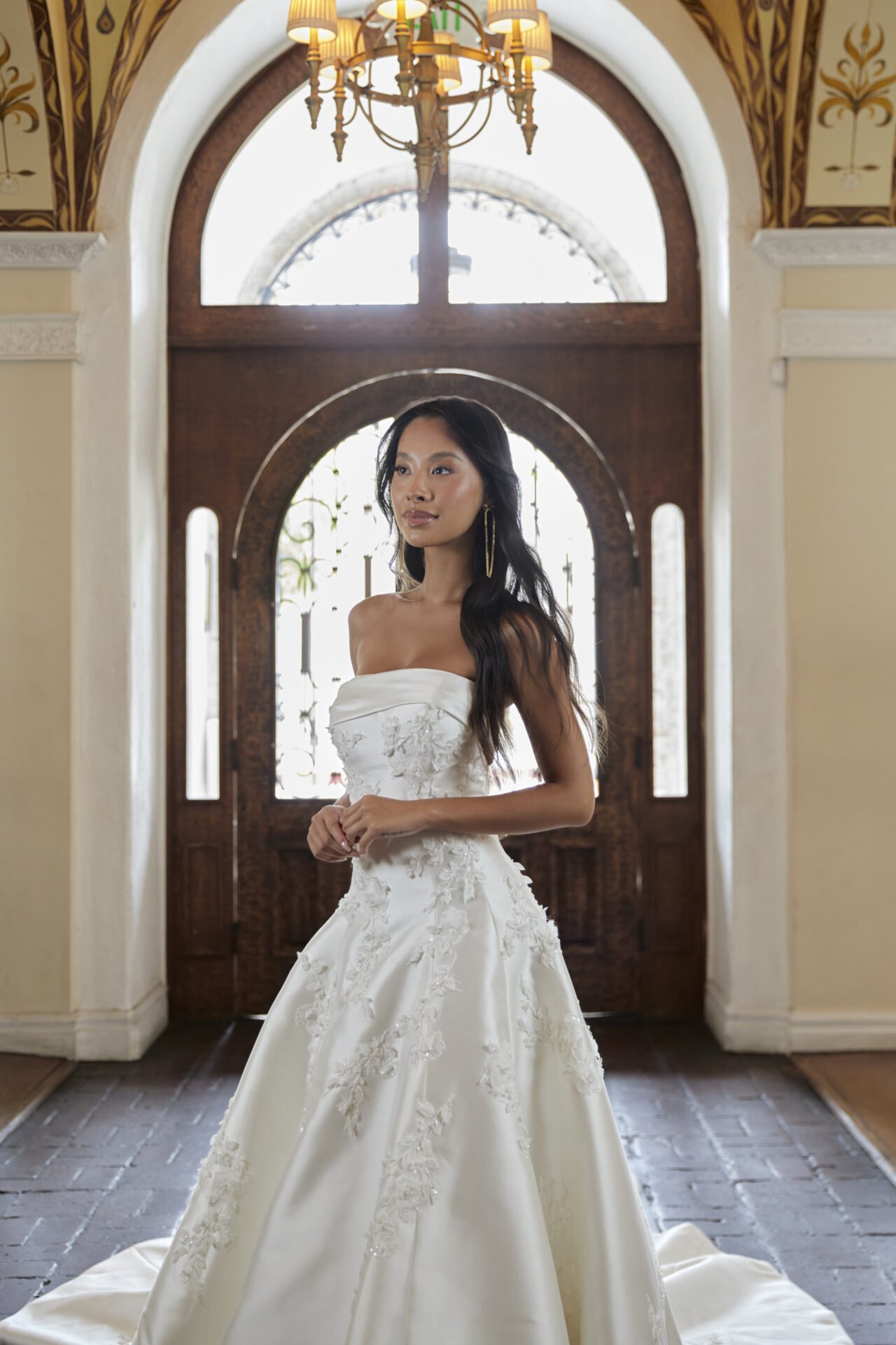 A woman in a white wedding dress standing inside a decorated church with wooden doors, ornate ceiling, and chandelier.