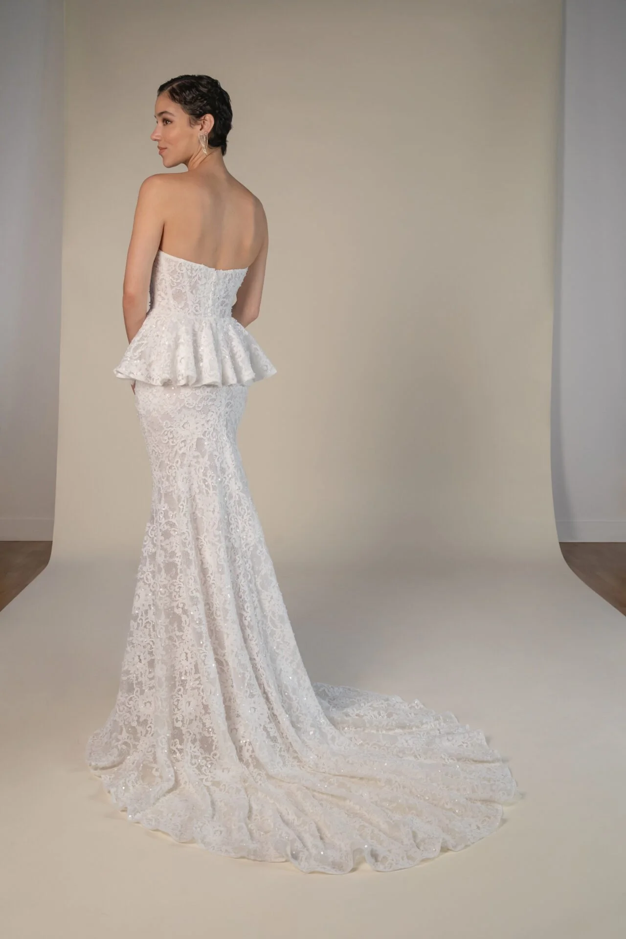A woman wearing an elegant white lace wedding dress with a fitted skirt, a peplum waist, and a strapless top, standing against a neutral background.