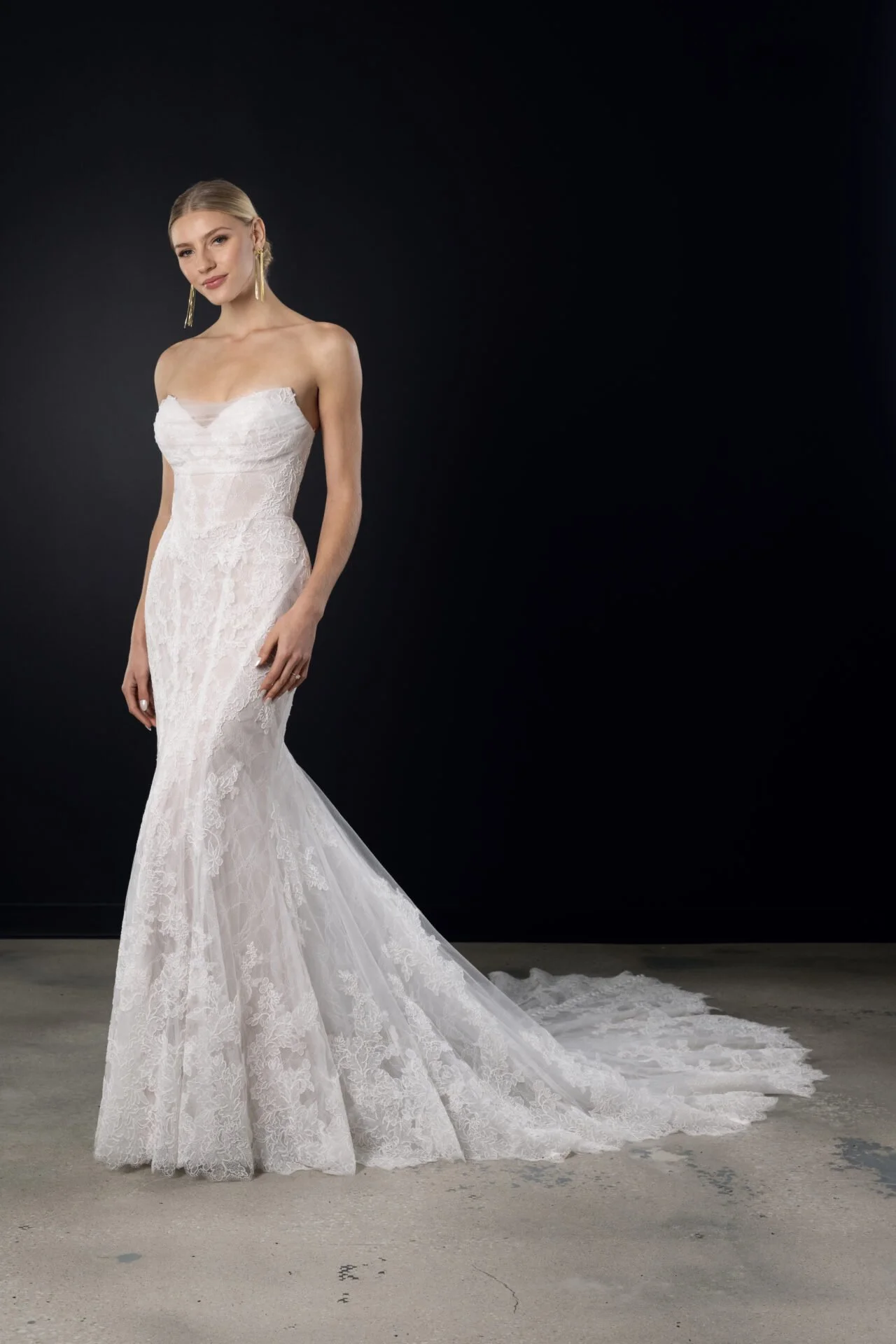A woman in a strapless, fitted white lace wedding gown with a long train, posing against a dark background.