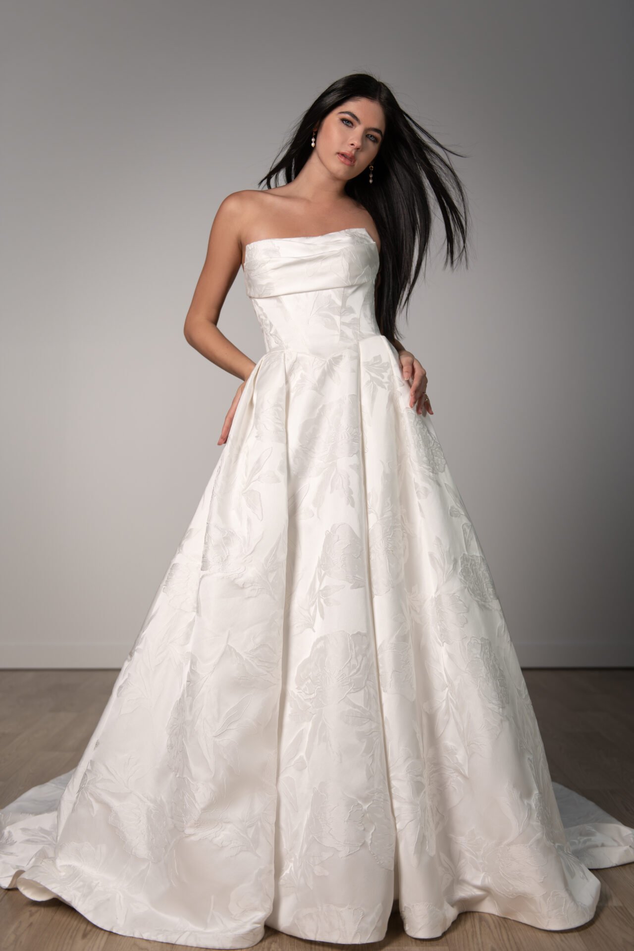 A woman wearing a strapless, white floral wedding gown with a full skirt, standing on a wooden floor against a plain gray background.