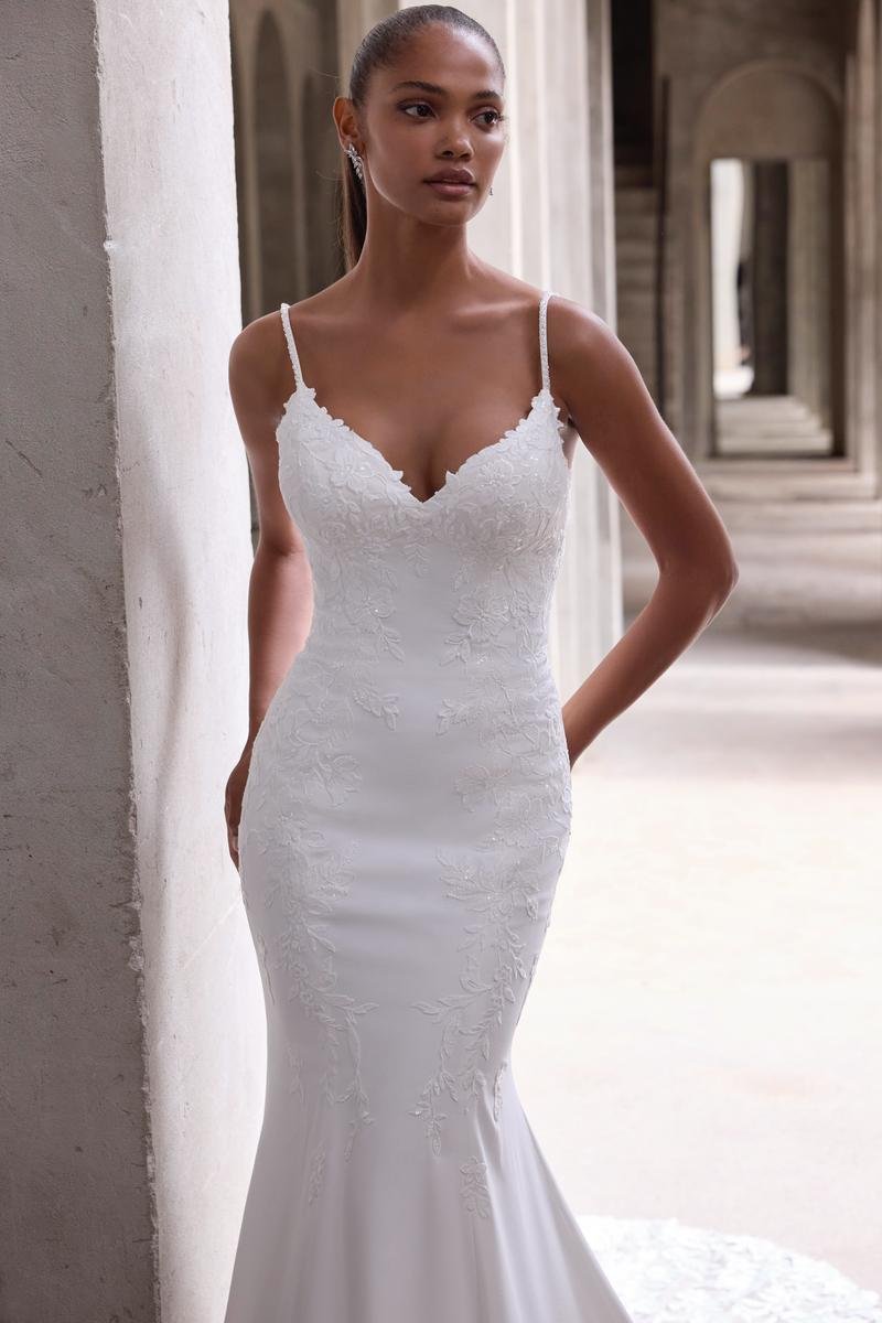 A woman in a white, form-fitting wedding dress with lace details, standing against a concrete wall in an architectural corridor.