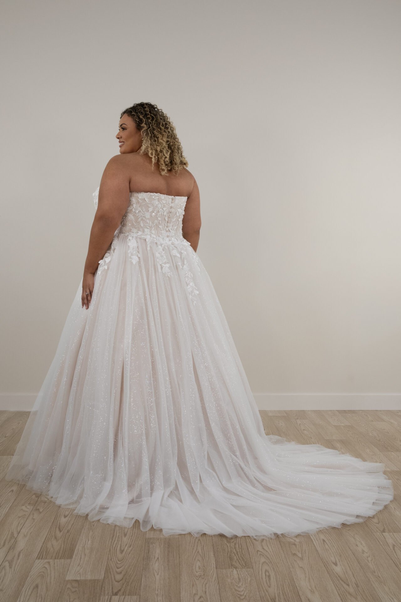 Woman in a white wedding gown with floral embroidery, standing against a plain wall and hardwood floor.