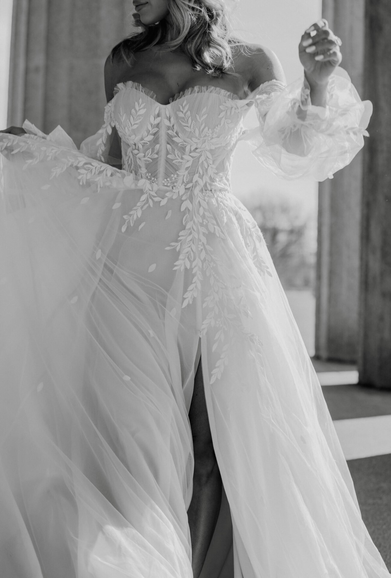 A woman wearing a wedding dress with lace detailing and sheer sleeves, lifting the skirt to reveal a thigh-high slit. The image is in black and white.