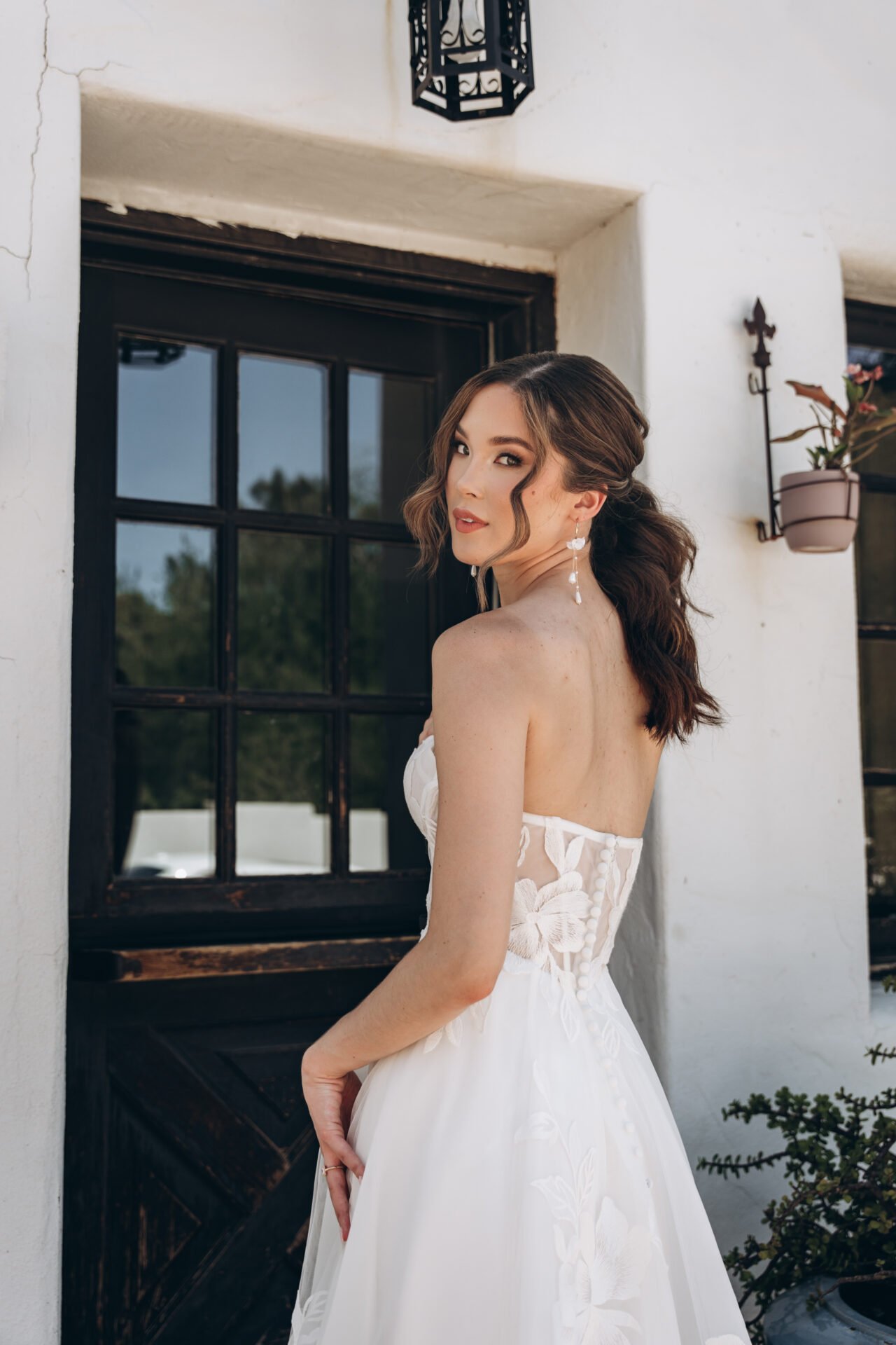 A woman in a white wedding dress with floral lace details, standing outside near a black-framed window, with brown hair styled in loose waves and wearing earrings.