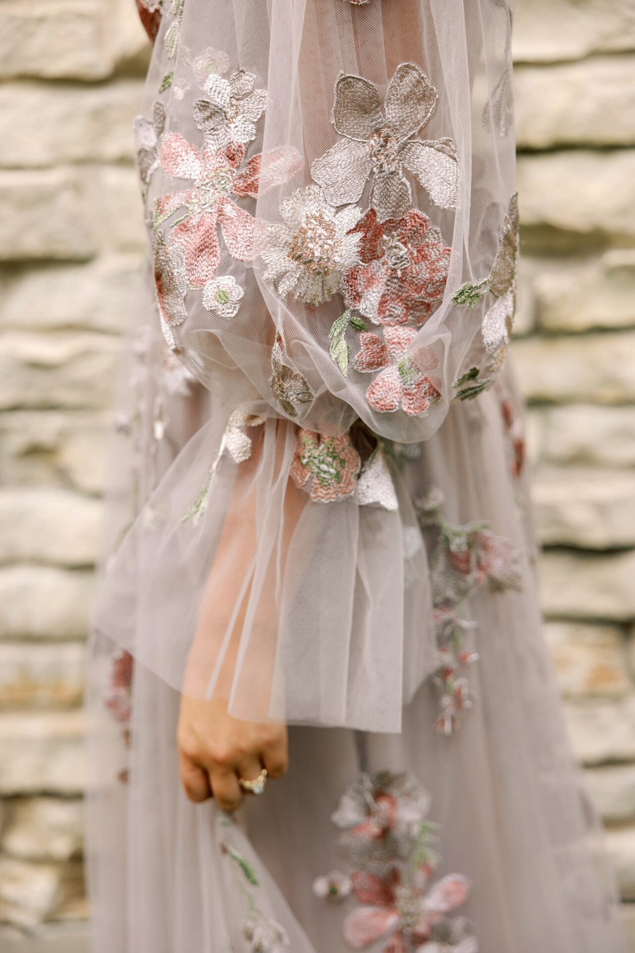 Close-up of a sheer, embroidered dress with floral designs, featuring pink, silver, and green embroidered flowers and a person's hand with a ring.