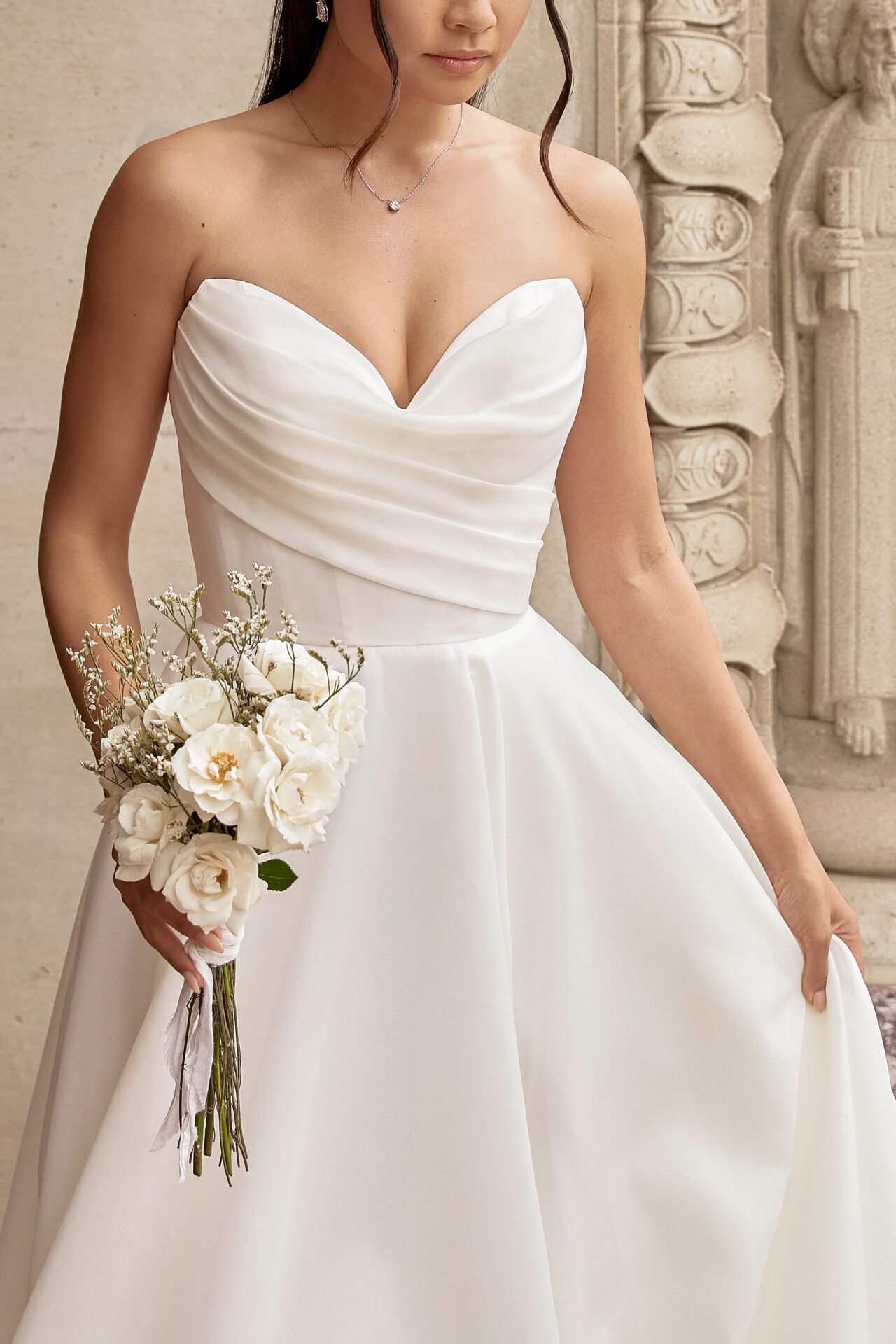 A woman wearing a strapless white wedding dress, holding a bouquet of white flowers, standing near carved stone architectural details.