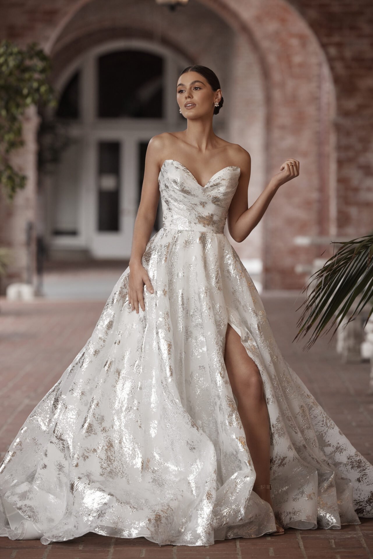 A woman wearing an elegant white and gold strapless wedding gown with a high slit, standing in front of a brick building and arched window.