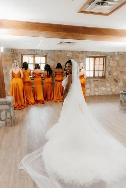 Bride in a white wedding dress with a long train and veil smiling at the camera, with five bridesmaids in orange dresses standing with their backs to the camera in a rustic room with stone walls and wooden beams.