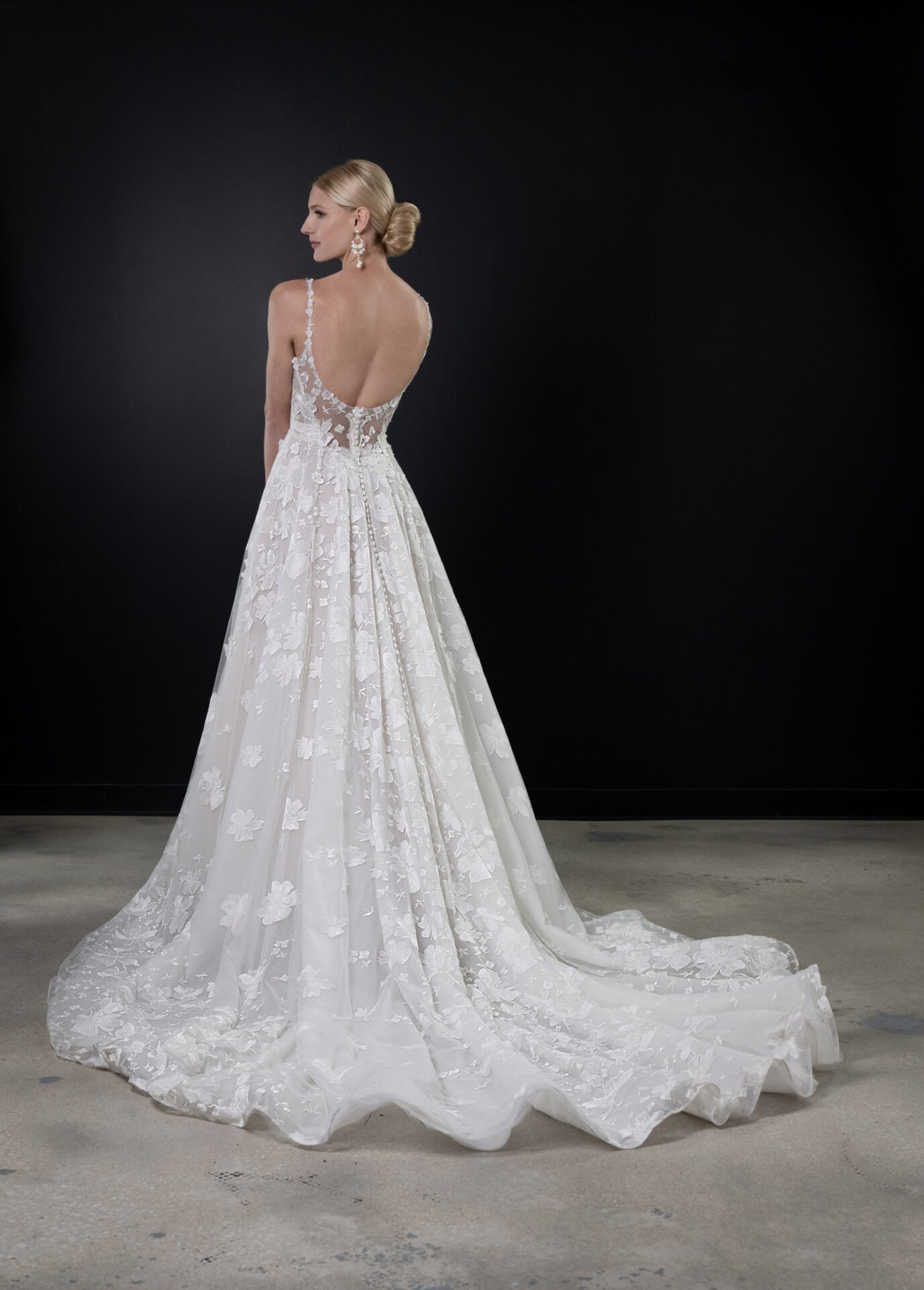 Woman standing in a wedding dress with lace floral details, open back, and train, against a dark background.