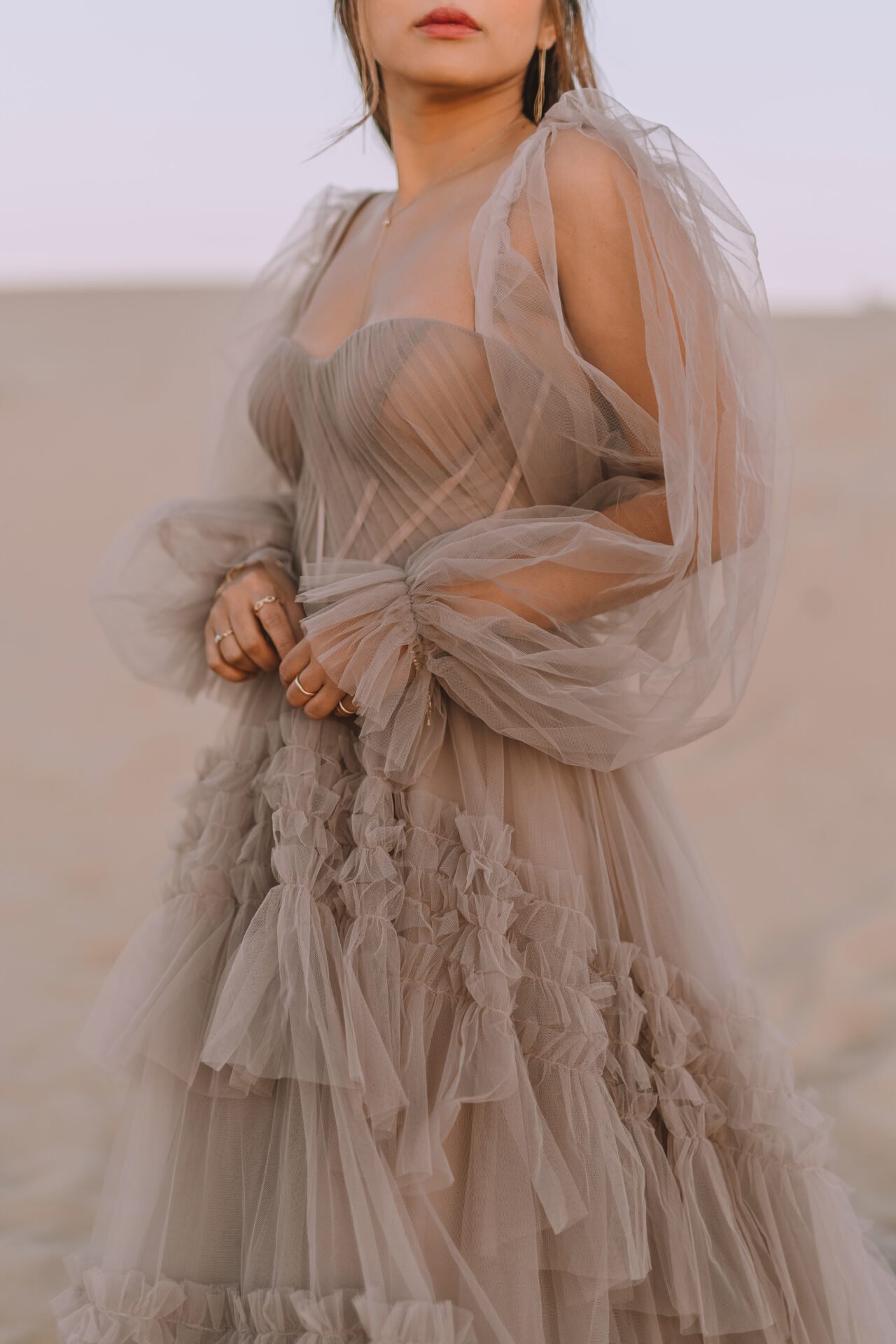 A woman wearing a beige, ruffled, sheer gown with puffed tulle sleeves, standing outdoors with a blurred neutral background.