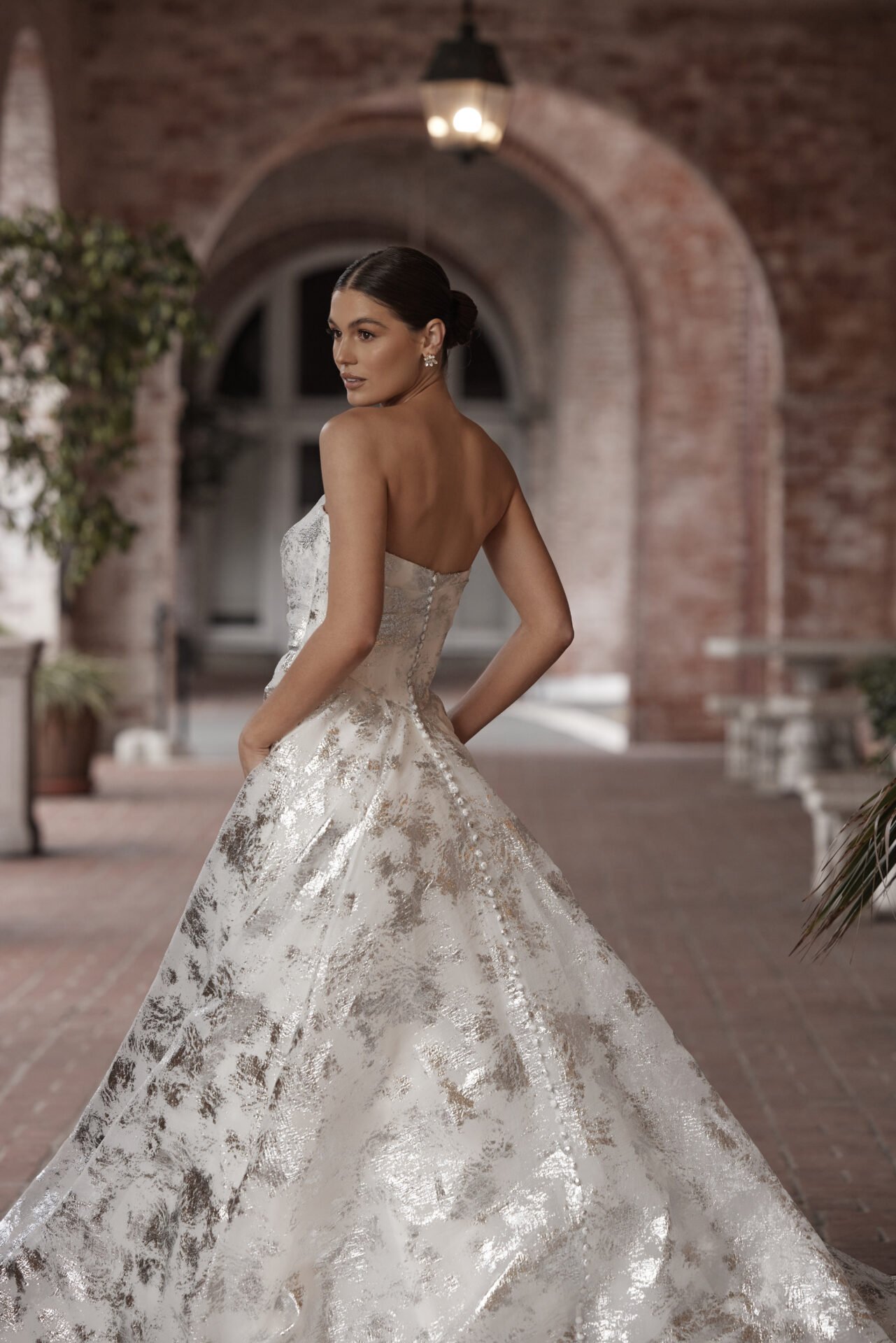 A woman in an elegant strapless wedding gown with metallic gold and silver patterns, standing outdoors under a brick archway with warm lighting.