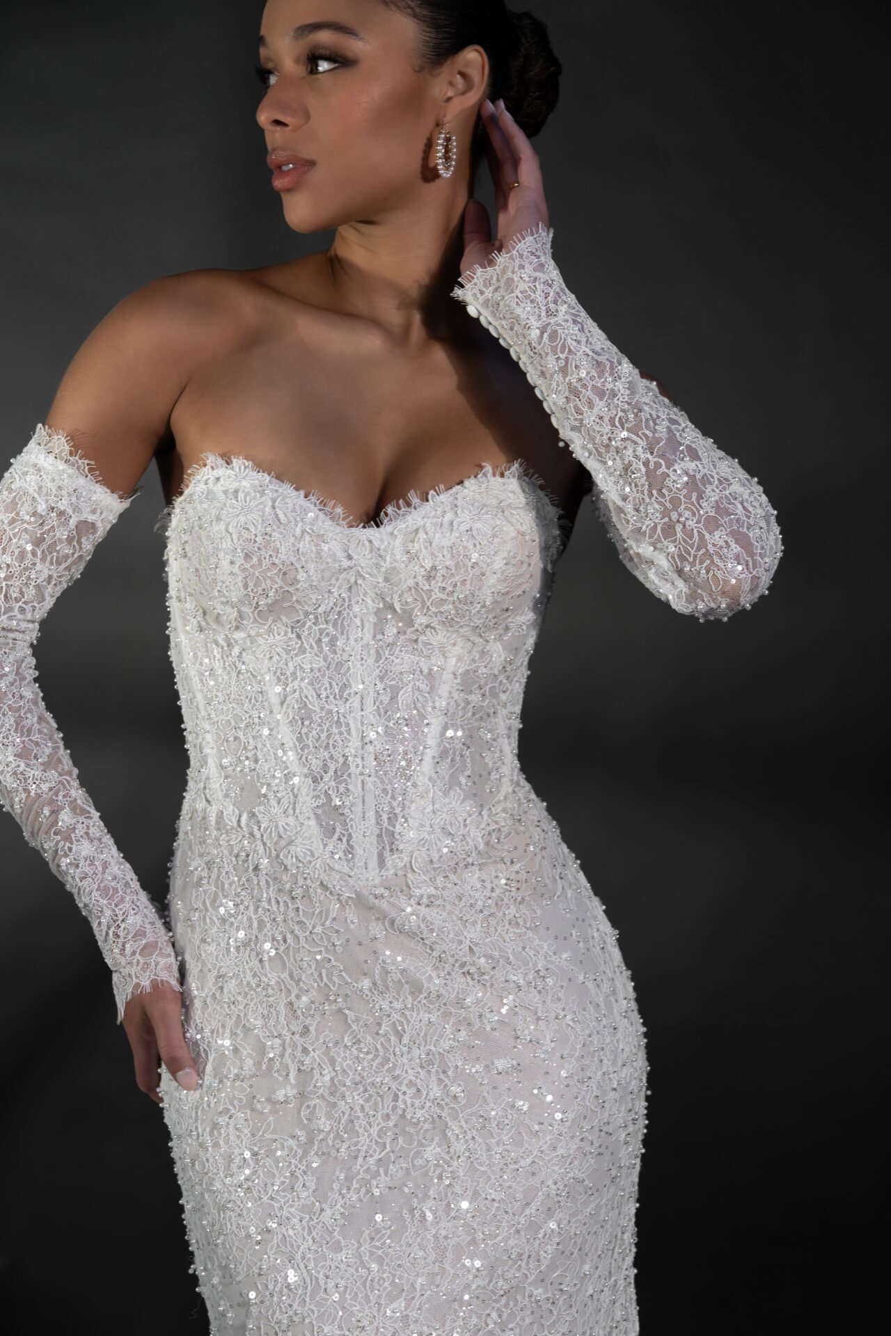 A woman wearing an elegant white wedding dress with intricate lace detailing and long lace sleeves. She has her hand near her ear and is wearing hoop earrings, with her hair styled in an updo. The background is dark.