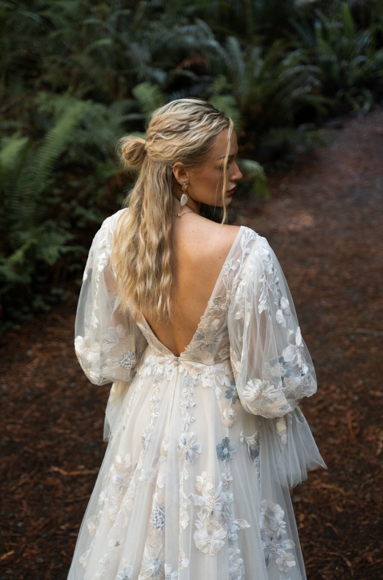 Woman in a white, floral embroidered dress with puffed sleeves and an open back standing on a dirt path surrounded by green foliage.