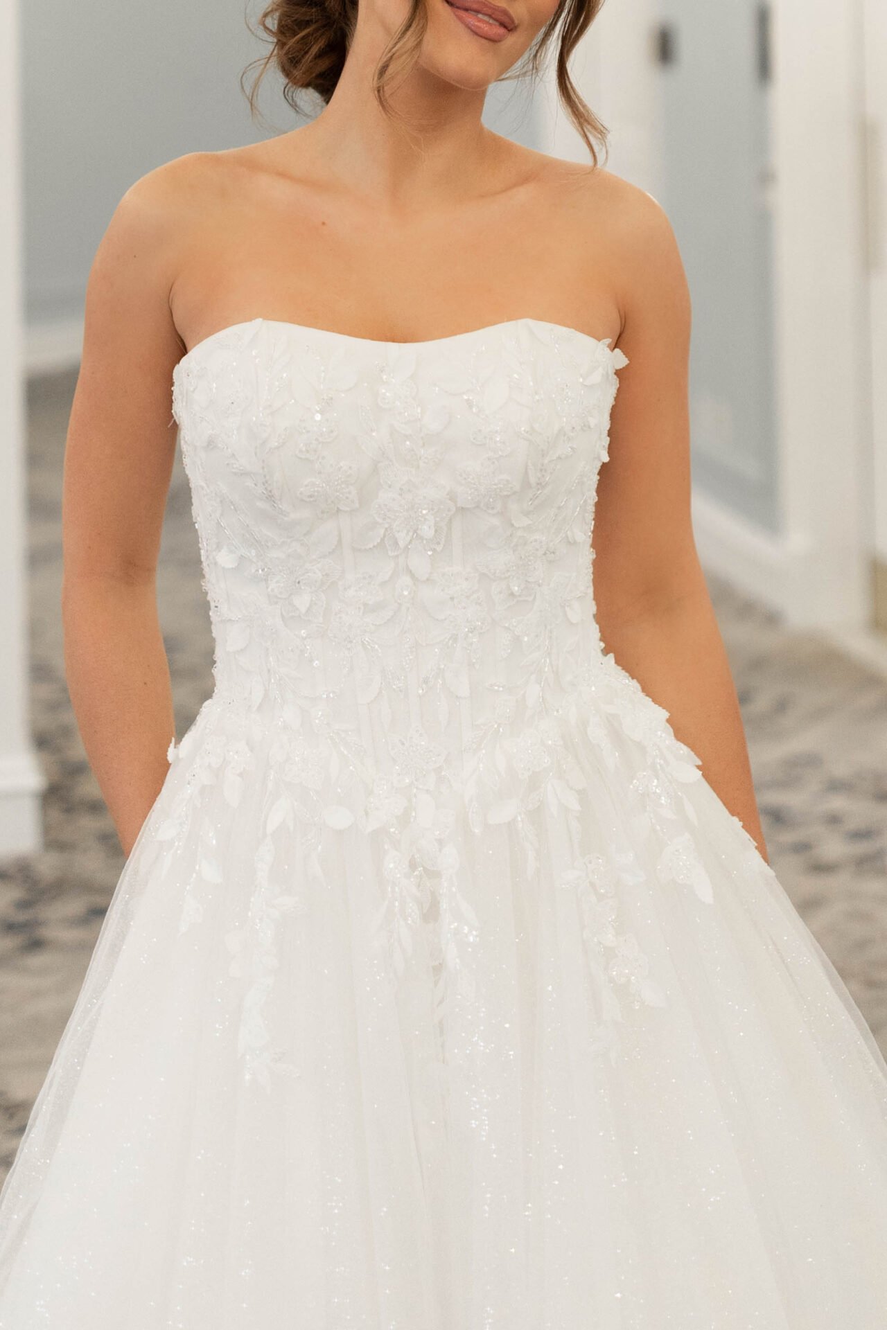 A woman wearing a white strapless wedding dress with floral embroidery, standing indoors in a well-lit room.