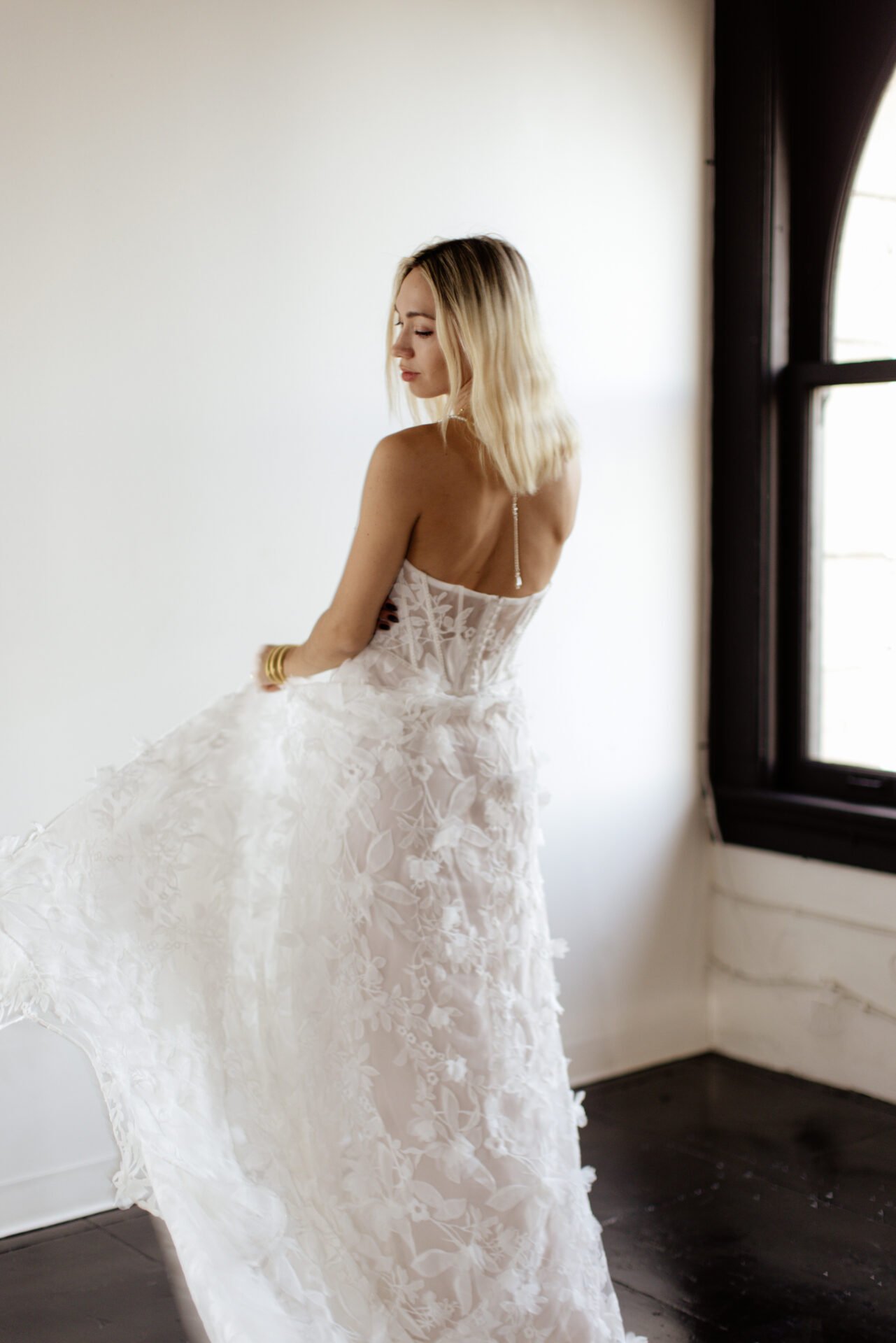 A woman with shoulder-length blonde hair wearing a white, floral embroidered wedding dress, standing near a window with black trim, looking down and slightly to the side.
