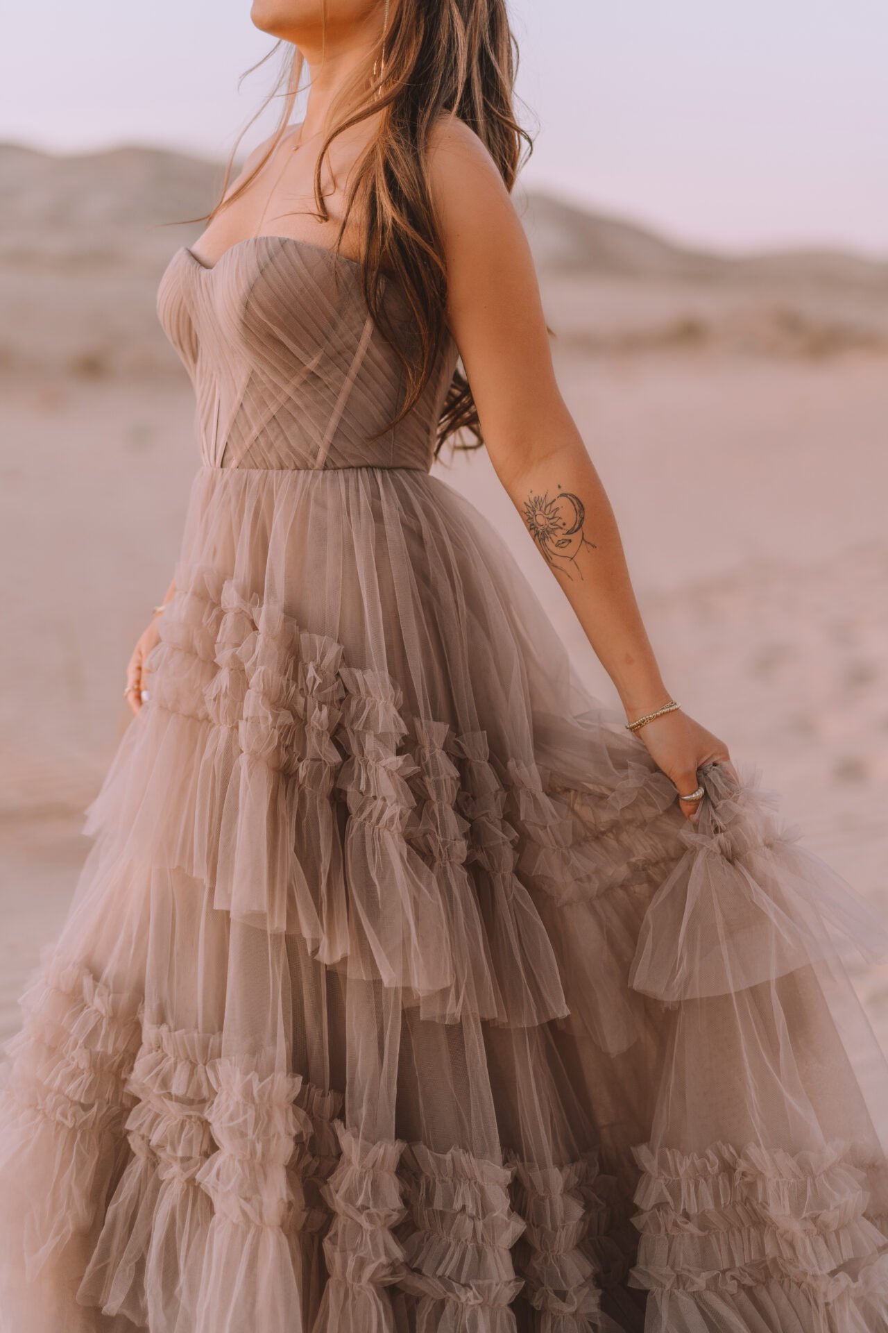 A woman in a layered, ruffled beige dress standing in a sandy desert landscape.
