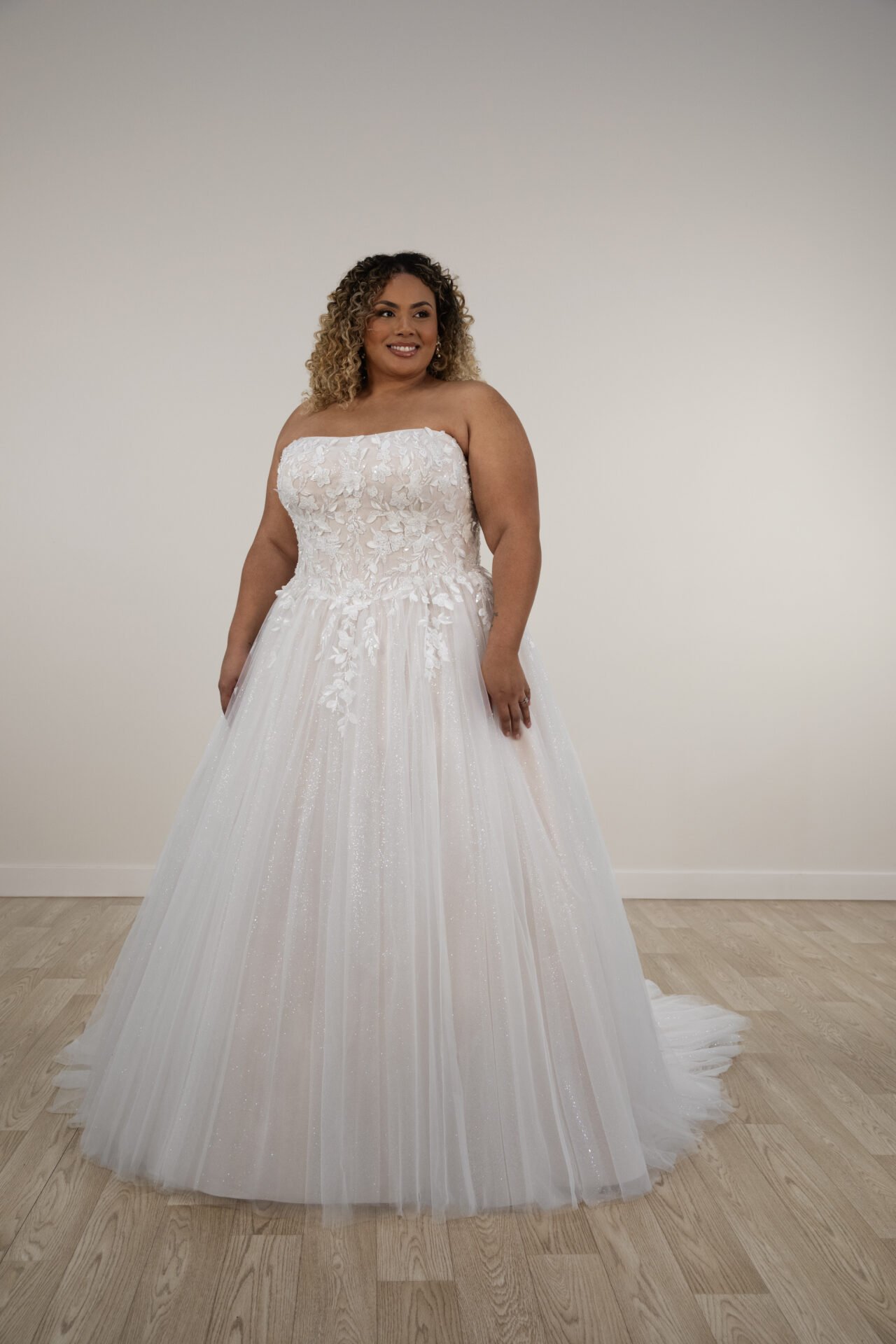 Woman in a strapless white wedding dress with floral lace detail on the bodice, standing against a plain off-white wall with light wood flooring.