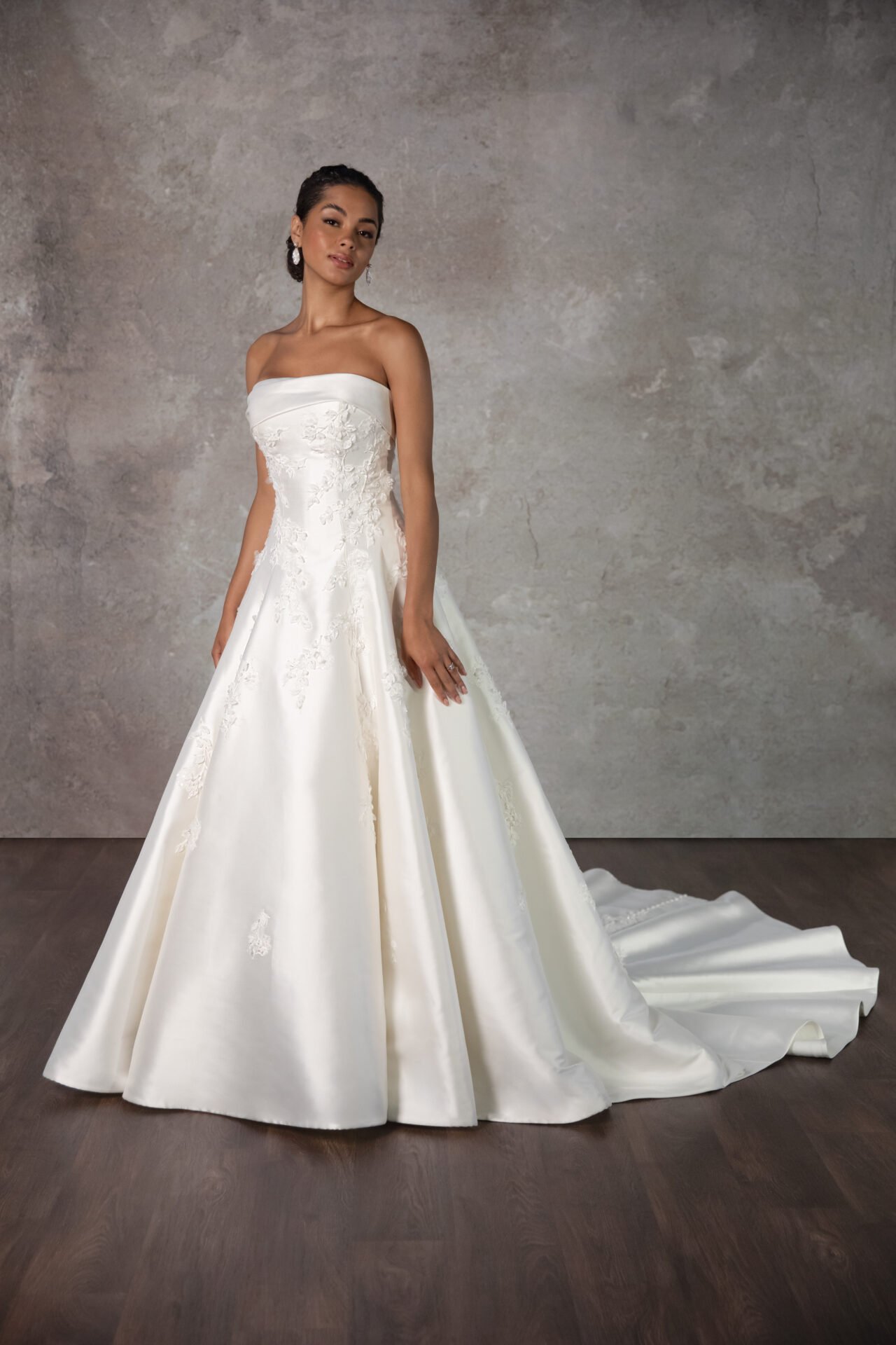 A woman wearing a strapless, white wedding gown with floral embroidery standing on a wooden floor against a textured wall.