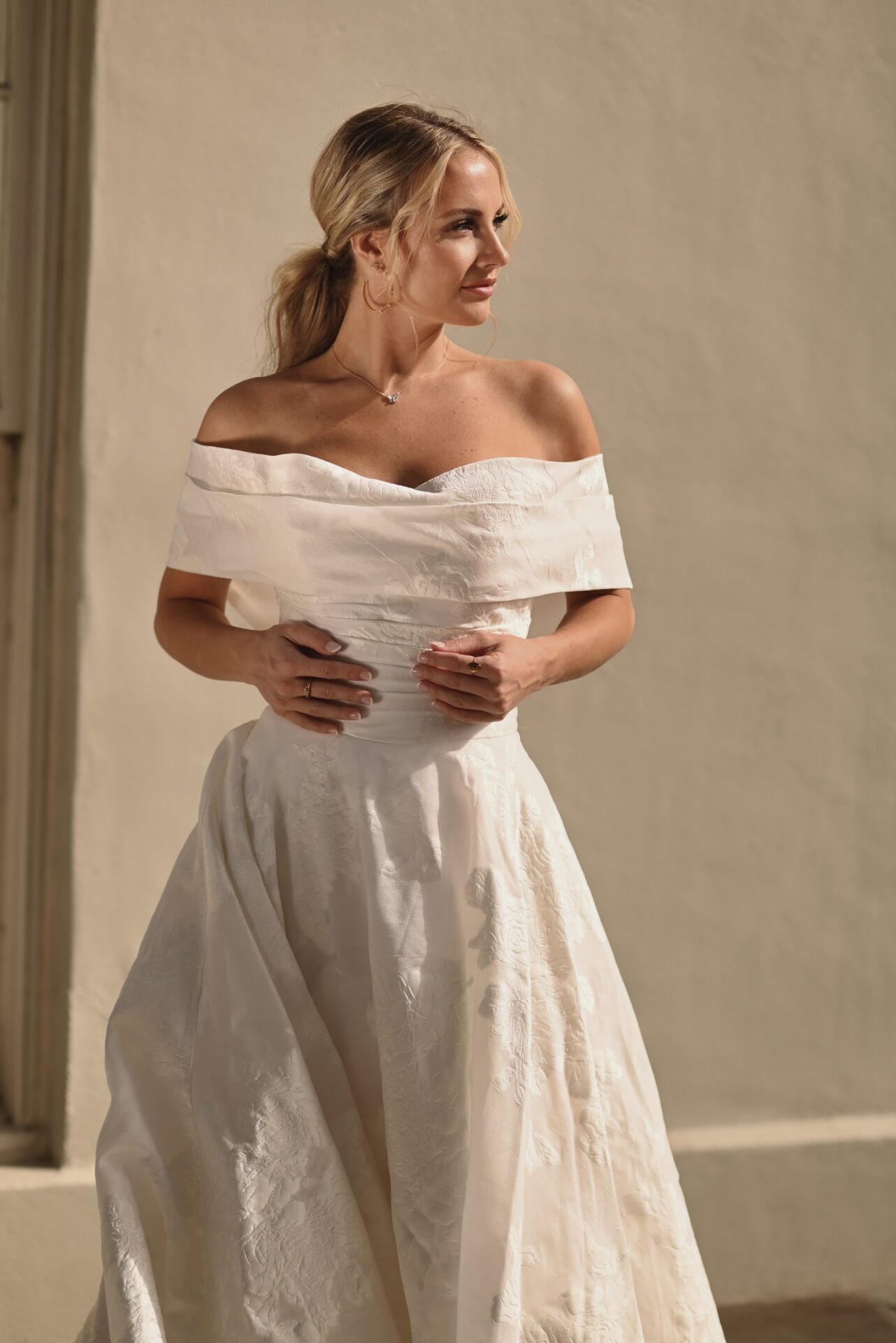 A woman in an off-shoulder white dress standing indoors against a neutral background, looking to the side.