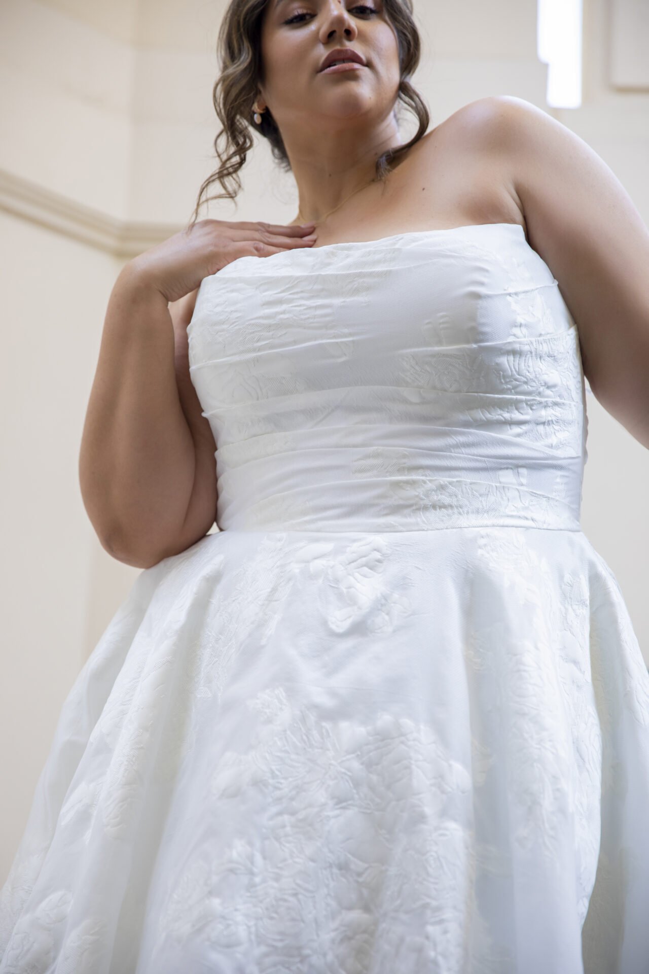 A woman wearing a white wedding dress, looking slightly downward, with her left hand resting on her shoulder.