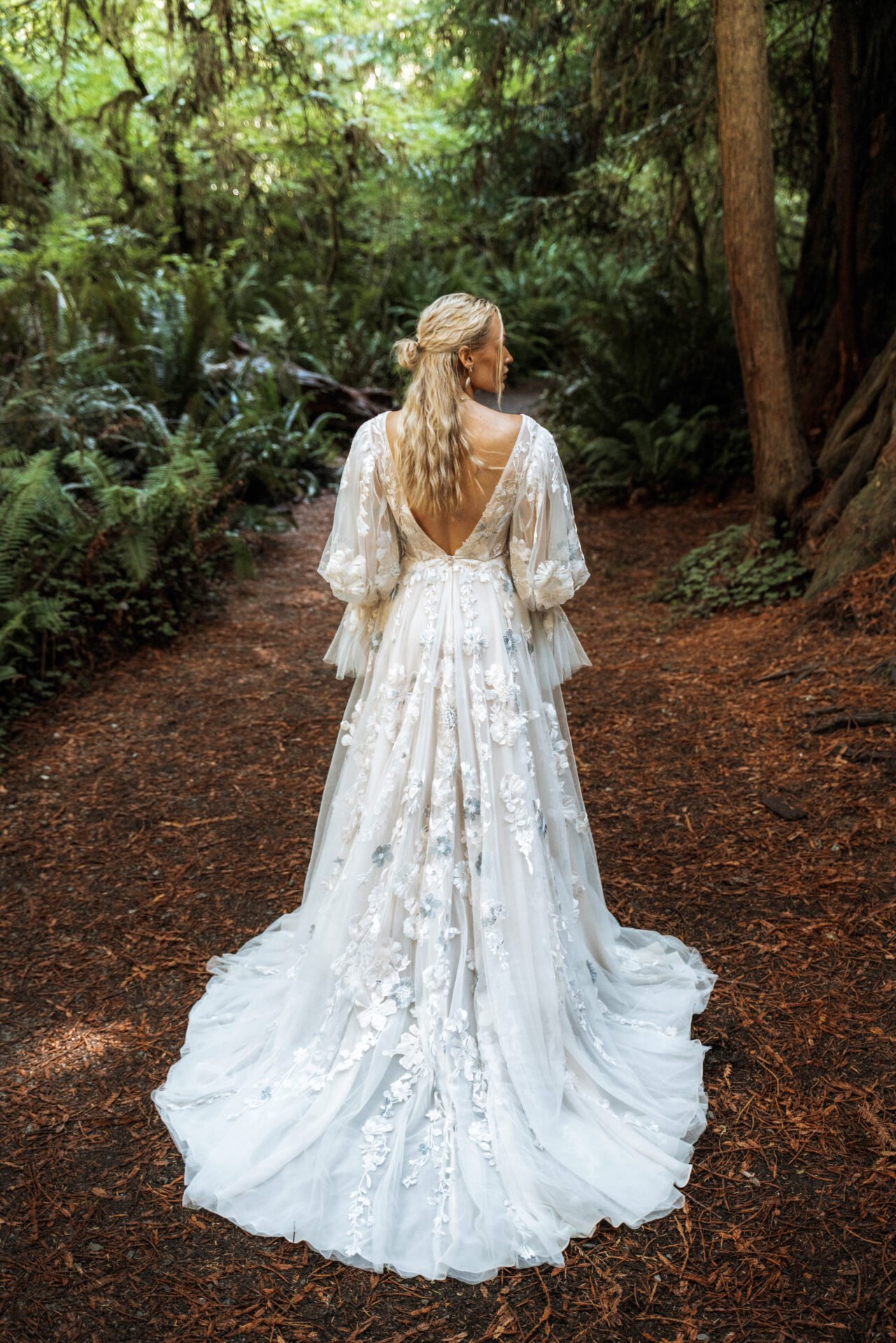 A woman in a white lace wedding dress with a low back walking along a forest trail surrounded by green trees and foliage.