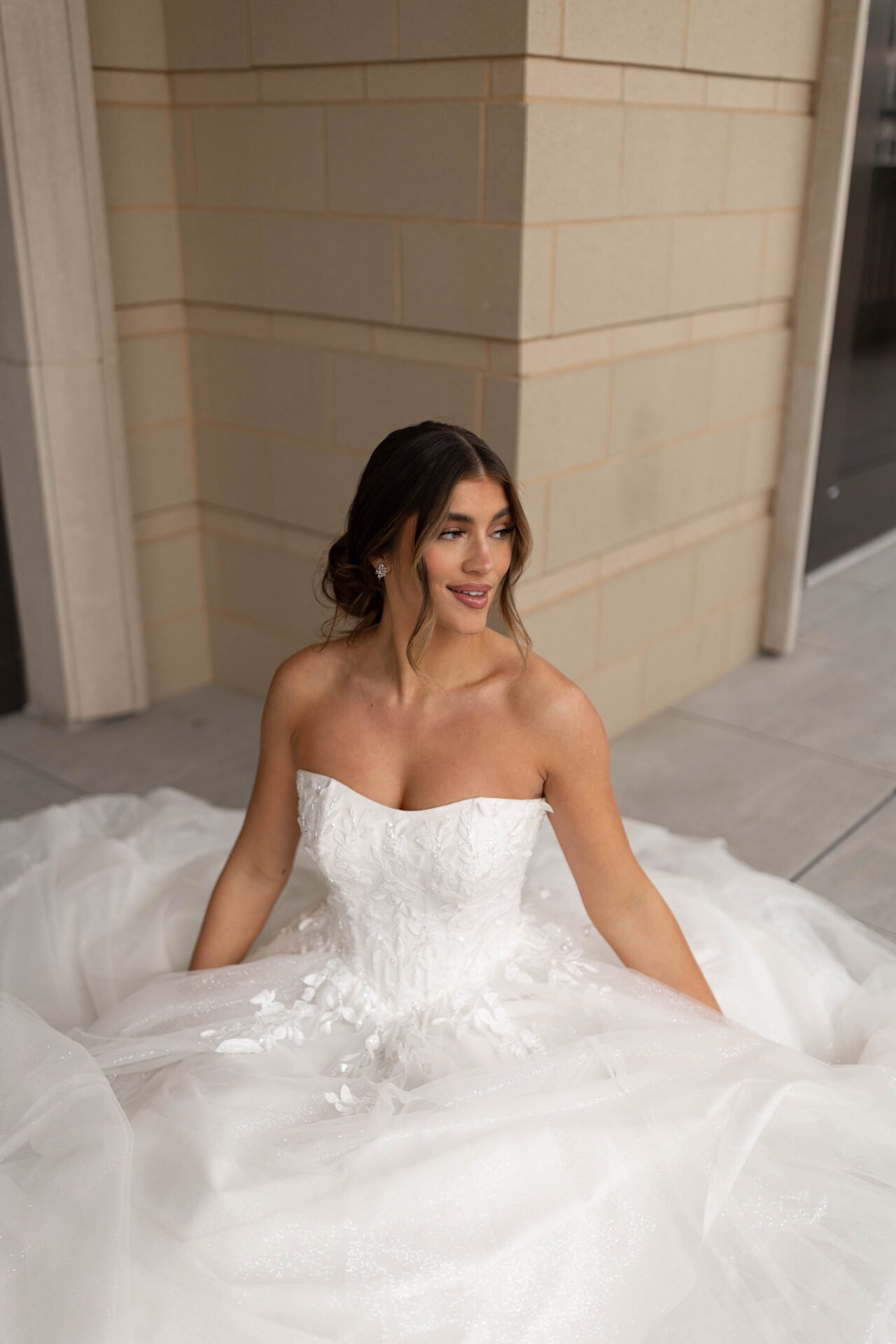 A woman in a strapless white wedding gown sitting on the floor near a beige brick wall, smiling and looking to her right.