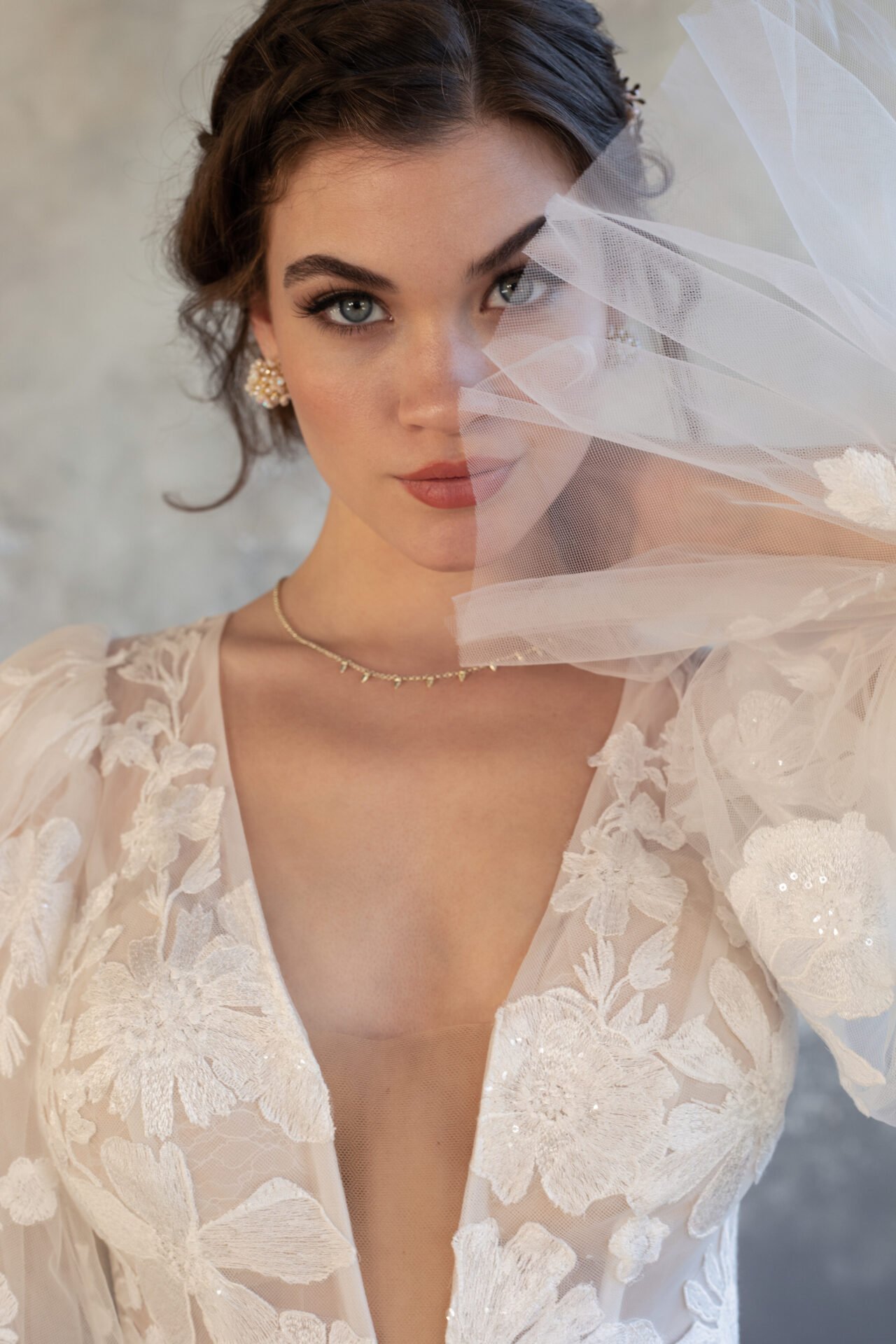 A woman dressed in a white bridal gown with embroidered floral details, partially covering her face with sheer tulle fabric, has wavy brown hair styled in an updo, and wears pearl earrings and a delicate necklace.