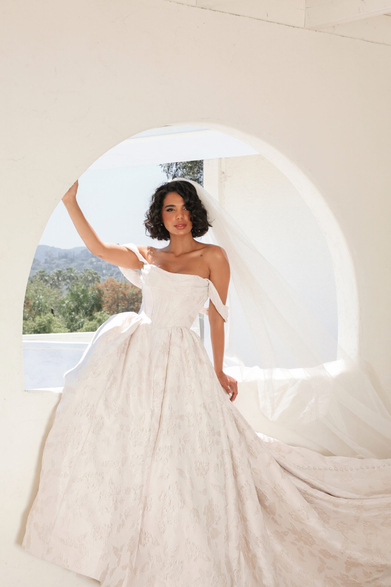 A woman in a white wedding gown standing inside near a circular window with a scenic outdoor view.