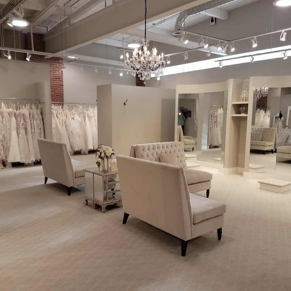 A bridal boutique with wedding dresses on racks, a chandelier, vintage-style seating, and large mirrors.