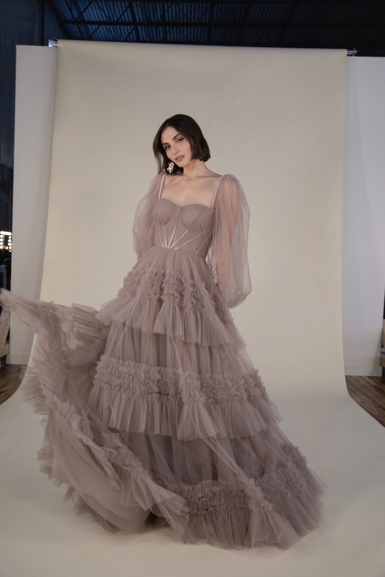 A woman in a flowing, layered, beige tulle dress with puff sleeves, posing in front of a neutral backdrop.