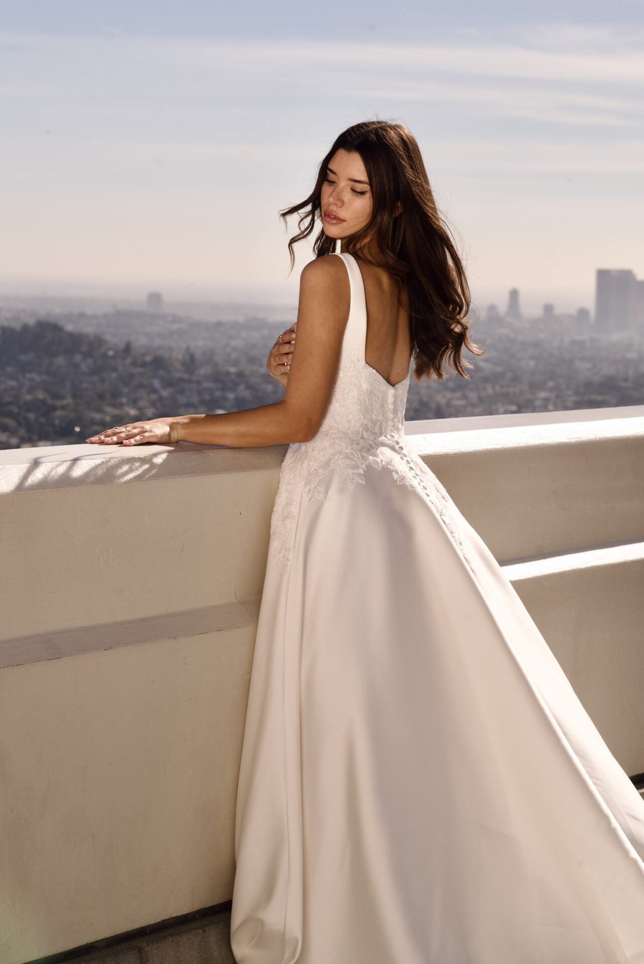 A woman in a white wedding dress standing on a balcony with a cityscape in the background, looking down with her eyes closed.
