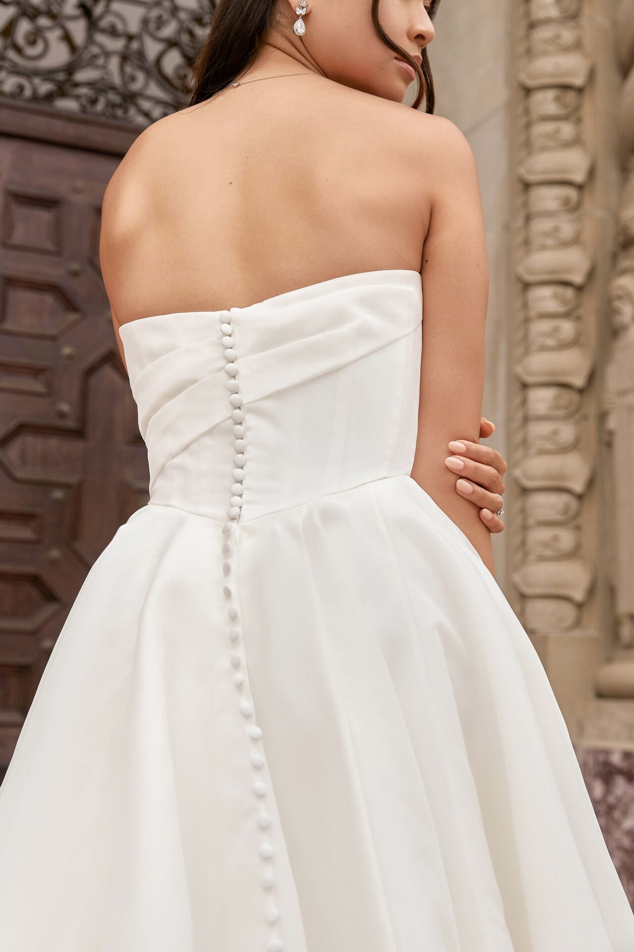 Close-up of a woman in a strapless white wedding gown with buttons down the back, standing indoors with carved wood and stone architecture in the background.