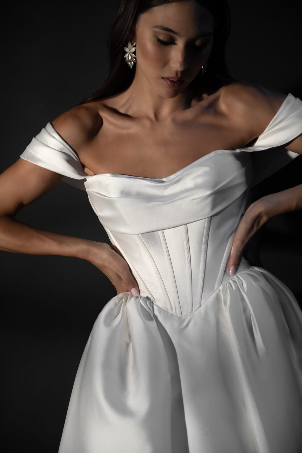A woman wearing a white satin off-shoulder dress with a fitted corset, standing against a dark background.