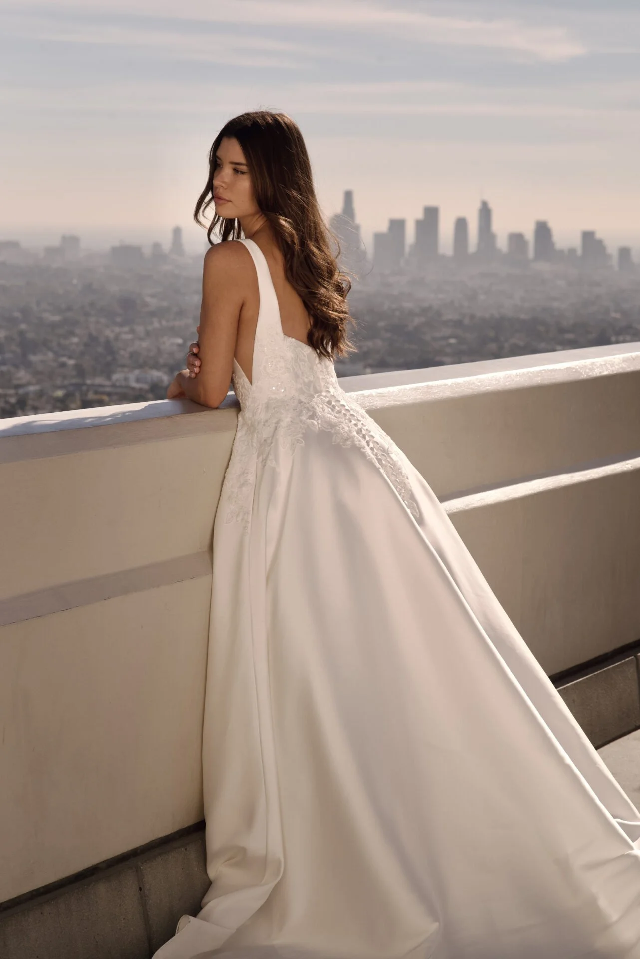 A woman in a white wedding dress with lace details stands on a balcony overlooking a city skyline during sunset. She has long, wavy brown hair and is resting her arms on the balcony railing, gazing thoughtfully into the distance.