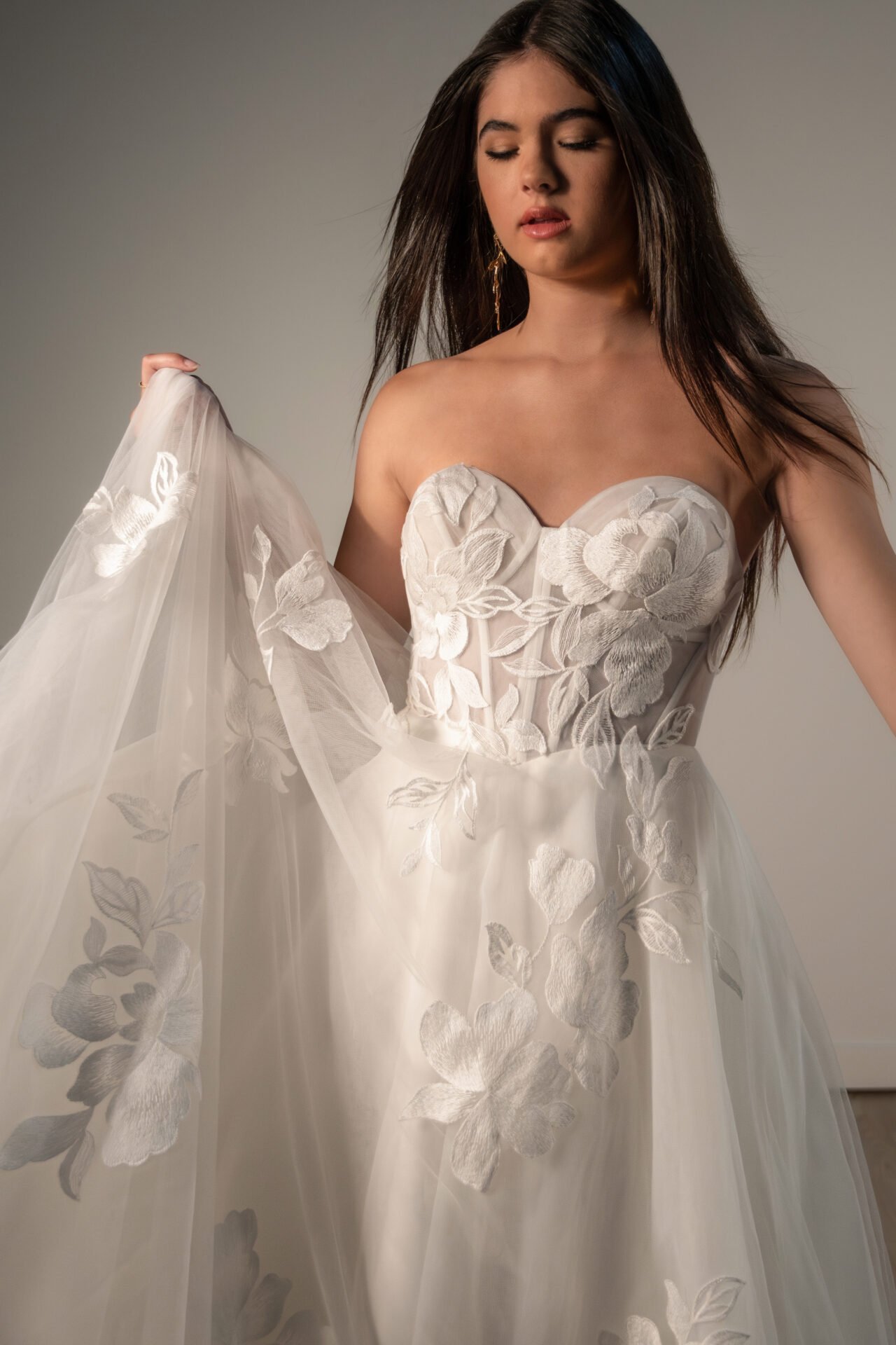 A woman in a strapless wedding dress with floral embroidery, holding the veil and looking down with closed eyes.