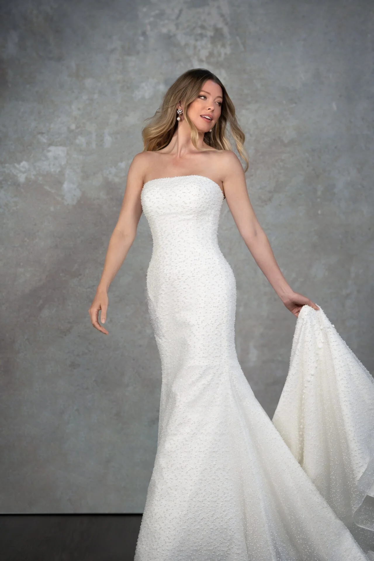 A woman modeling a strapless white wedding dress with a fitted bodice and flowing skirt, standing against a textured gray wall.