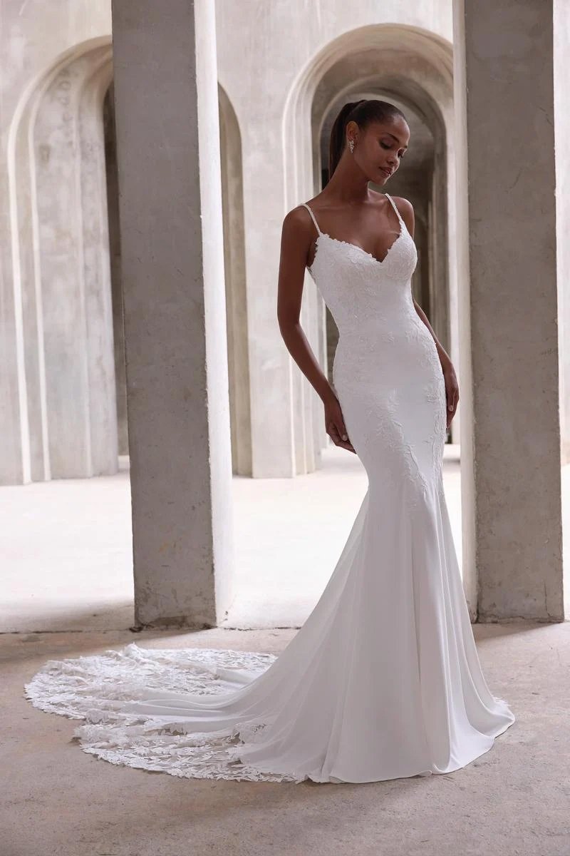 A woman in a white wedding dress with thin straps, lace details, and a long train standing in a stone archway.