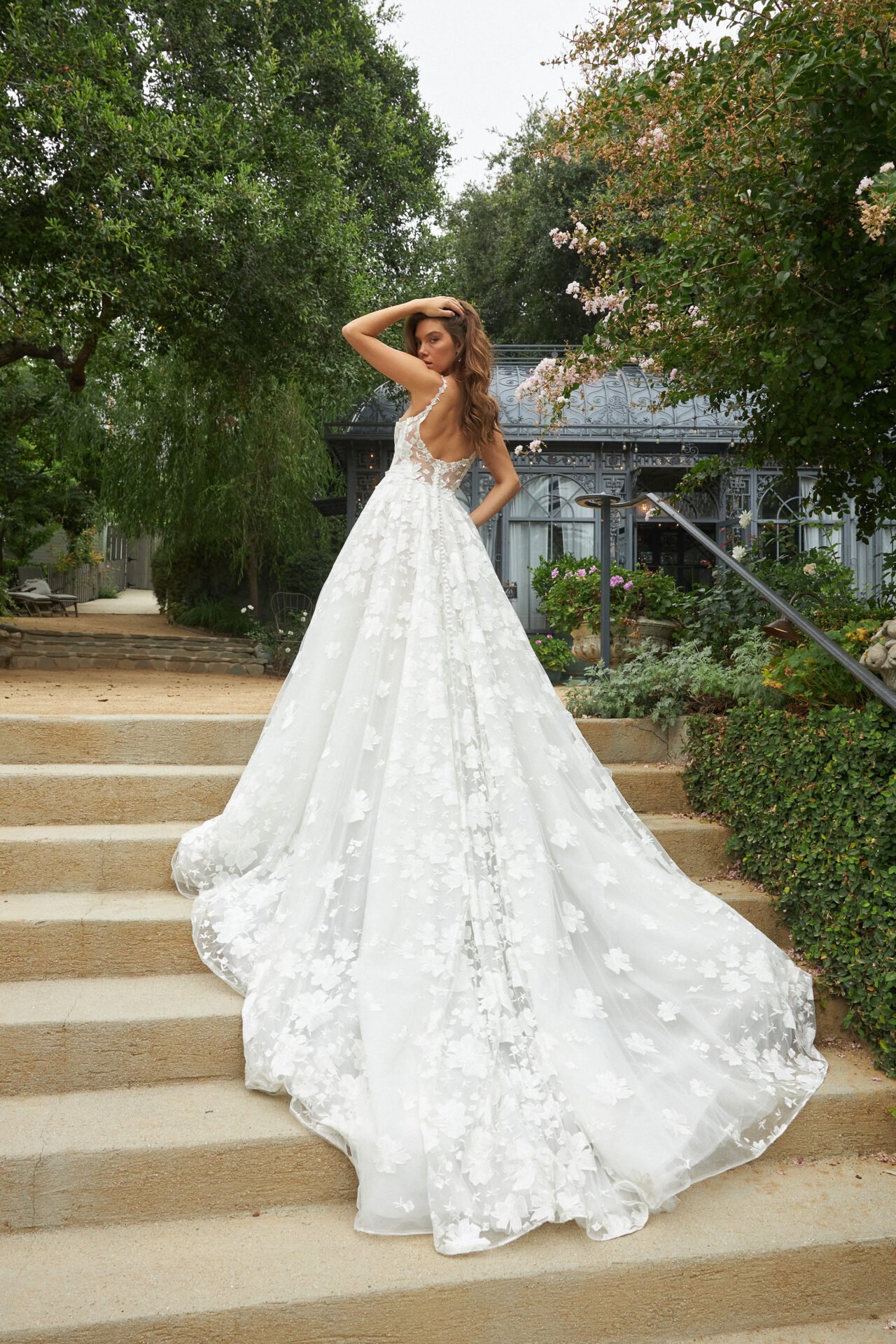 A woman in a white wedding dress with floral lace details stands on stone steps outdoors, with greenery and a decorative glass greenhouse in the background.