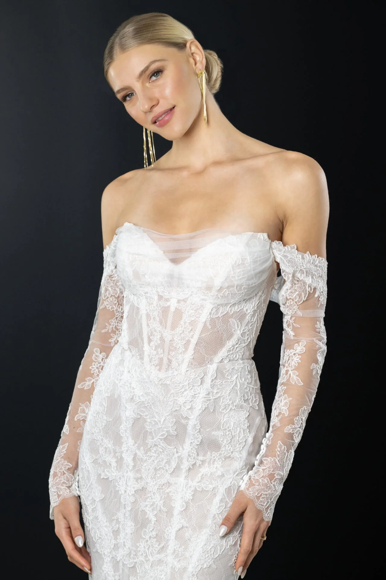 A woman wearing an off-shoulder white lace wedding dress with long sleeves and gold earrings, posing against a black background.