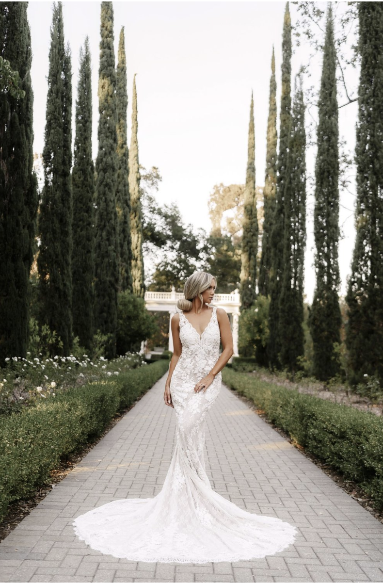 A woman in a white lace wedding dress with a train walking along a paved garden pathway lined with green shrubs and tall cypress trees, with a white balustrade visible in the background.