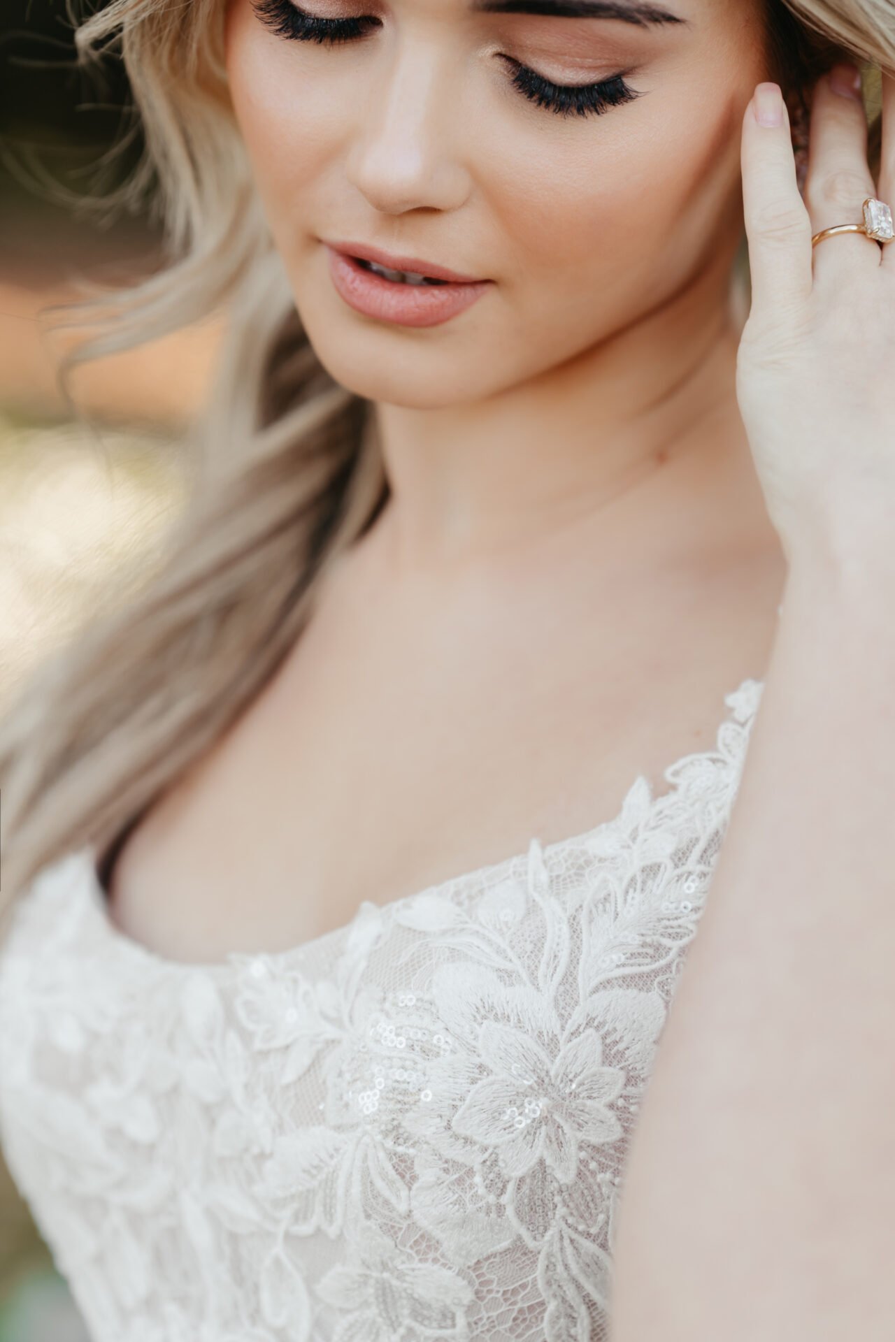 Close-up of a woman with blonde hair, wearing a white lace dress, with her eyes closed and hand touching her ear, outdoors.