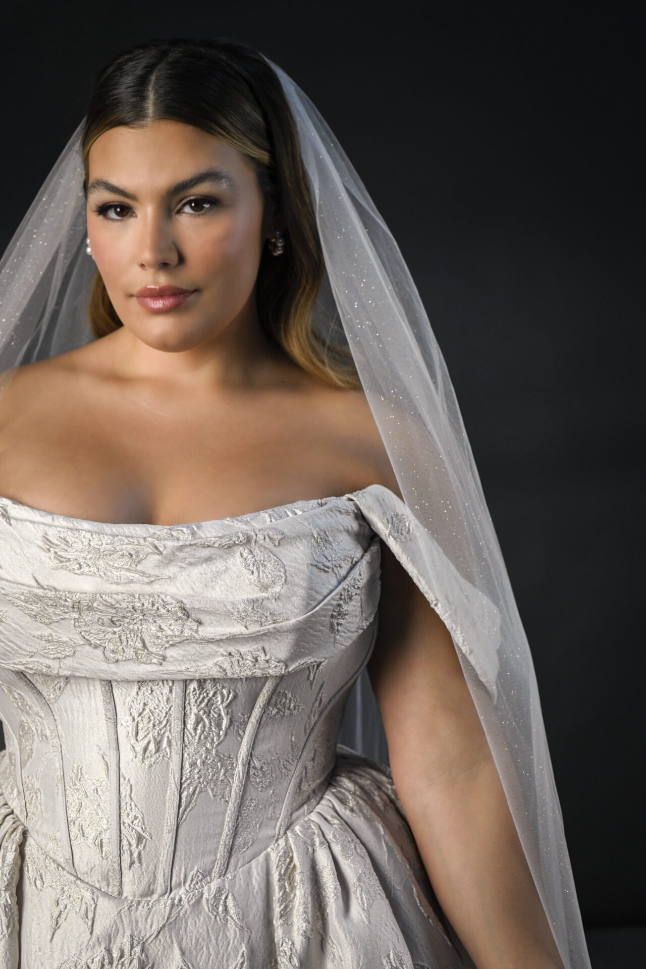A woman wearing a wedding dress with a veil, sitting against a dark background.
