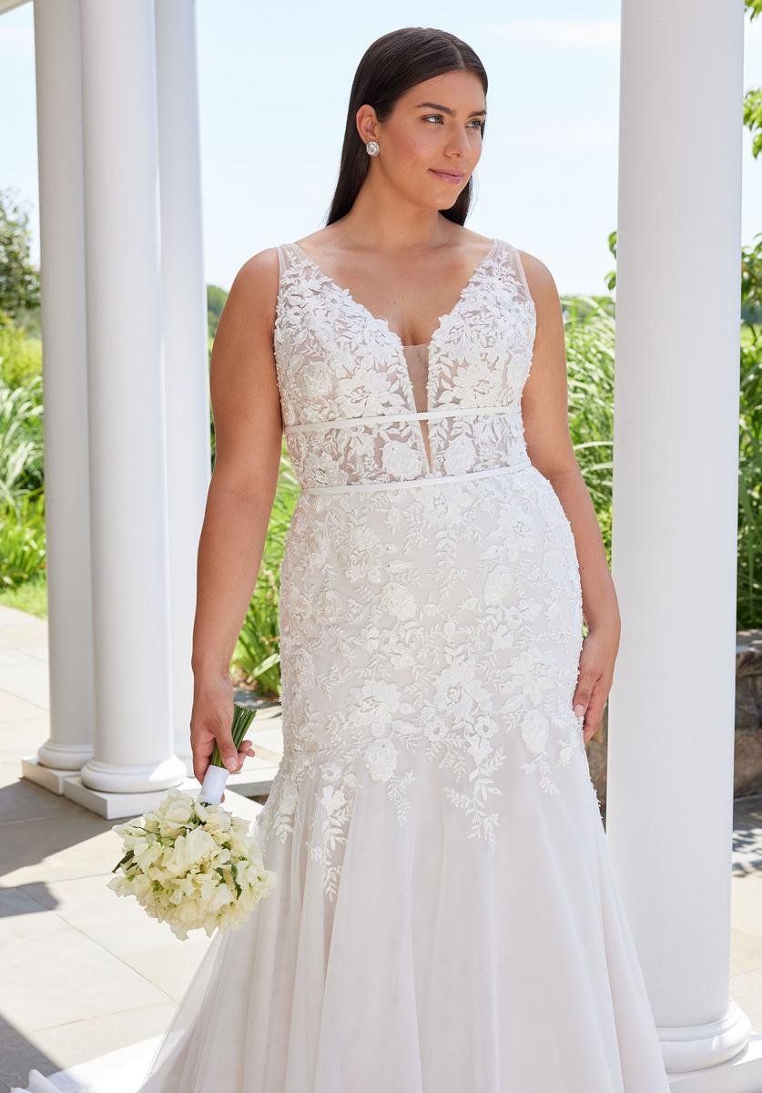 A woman in a white lace wedding gown holding a bouquet of white flowers, standing outdoors near white columns and greenery.
