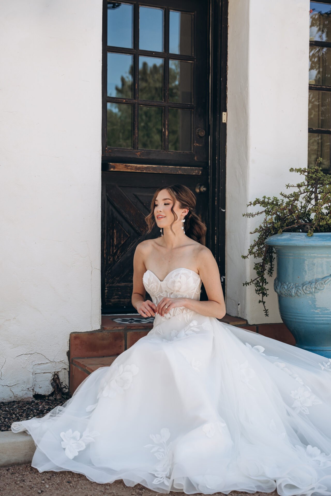 A woman in a strapless white wedding dress with floral embroidery sits on brick steps outside a white building with a black door. There is a large blue planter with green plants beside her.