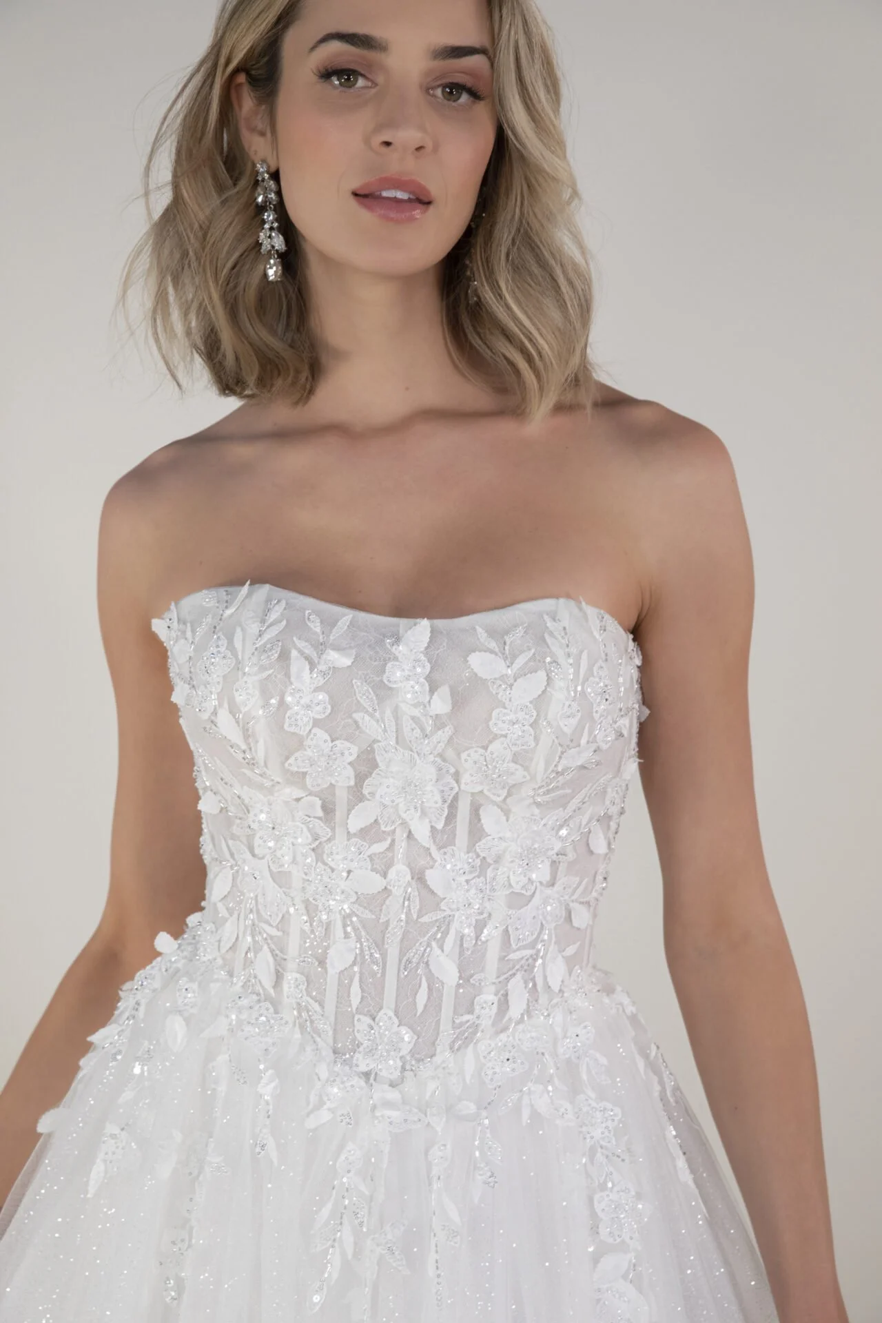 A woman wearing a strapless white wedding dress with floral lace details and embroidery, standing against a plain background.
