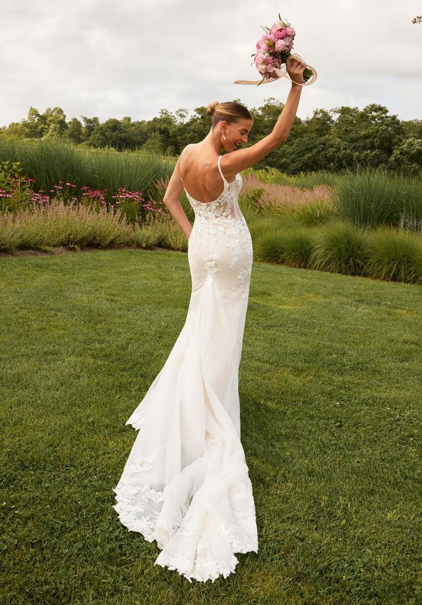 A woman in a white wedding gown holding a bouquet of pink flowers in the air outside on the grass with greenery and trees in the background.