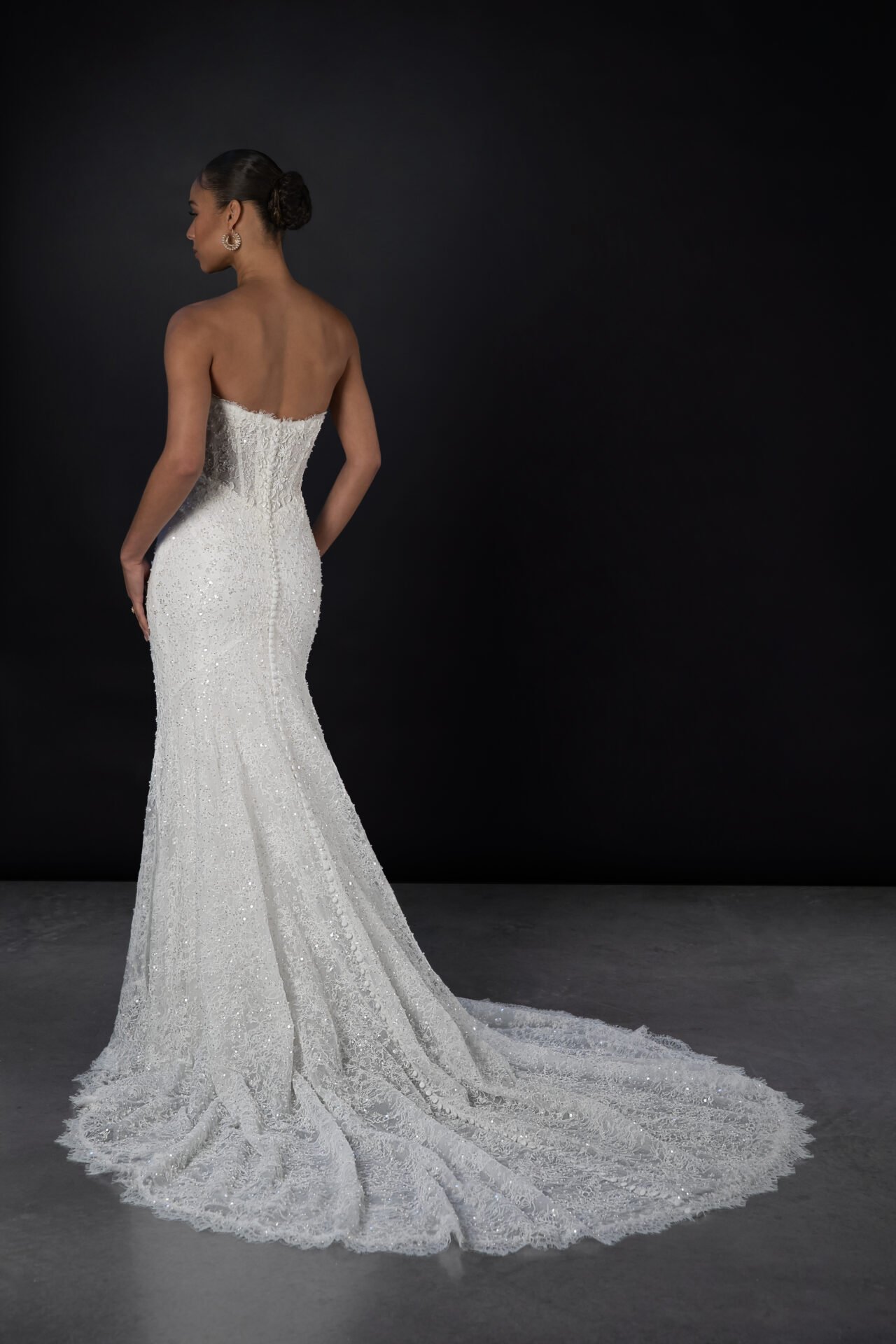 A woman in an elegant white strapless wedding dress with a train, standing against a dark background. The dress is adorned with intricate beading and embroidery.